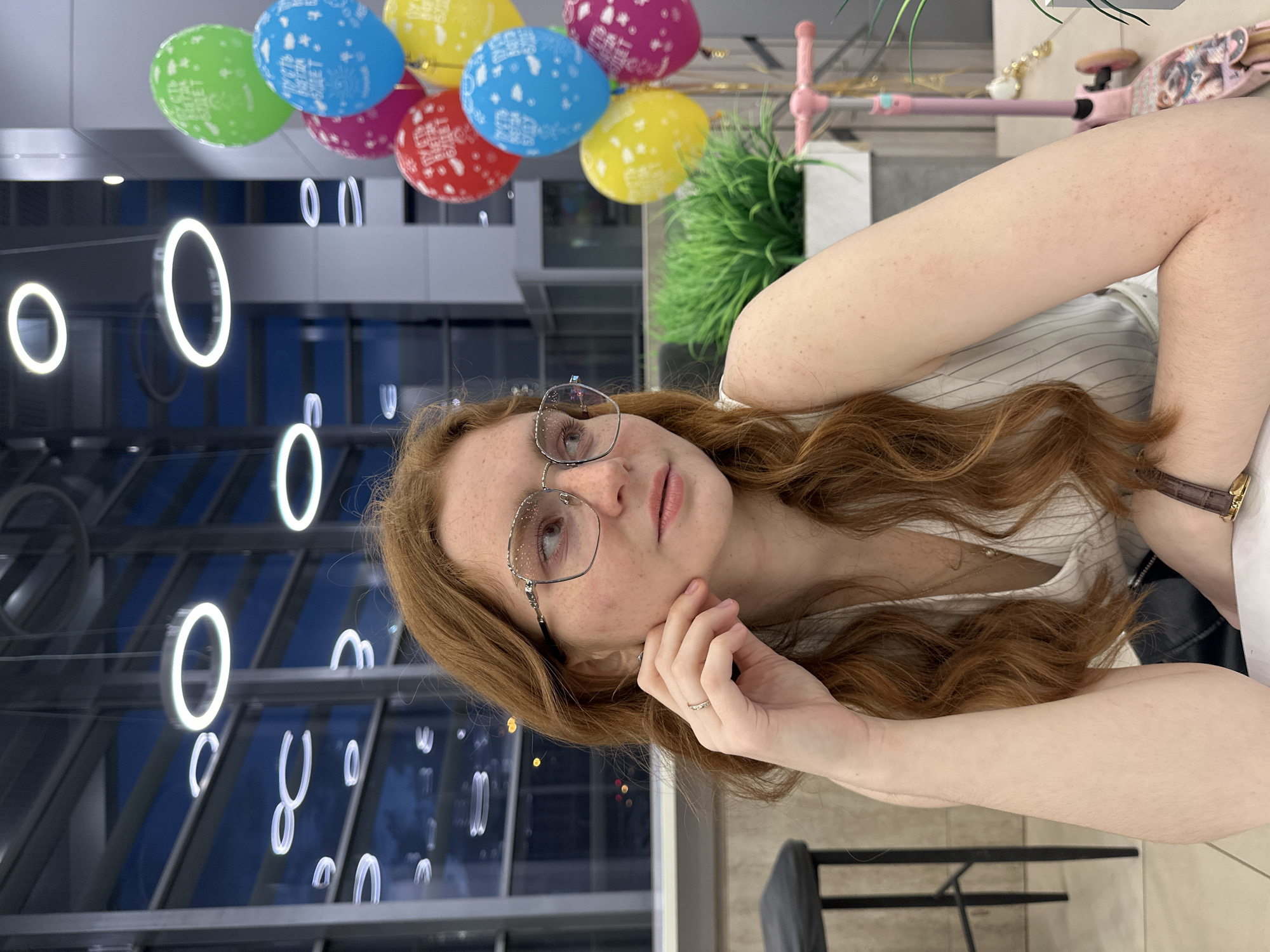 A young woman with long, wavy reddish-brown hair and visible freckles is pictured indoors, gazing thoughtfully upwards and to her right. She wears stylish glasses with decorative frames and a thin ring on her left ring finger, with her left hand resting gently on her jaw. Her right wrist displays a watch with a brown leather strap, and she is dressed in a light-colored, possibly striped, top.
The background suggests a modern, spacious interior, likely a commercial or public area in Kazan, Russia, characterized by large windows or reflective surfaces. Prominently featured are numerous glowing white circular light fixtures reflected in these surfaces. In the upper left, a vibrant cluster of balloons in green, blue, yellow, pink, and red indicates a celebratory event. Text such as "BEST WISHES" is visible on the blue balloons, and "GOOD LUCK" on the green ones. Hints of a green potted plant and a pink children's toy handle (possibly a scooter) are visible on a table to the right. The lighting suggests evening or nighttime. The overall scene conveys a calm, contemporary ambiance.