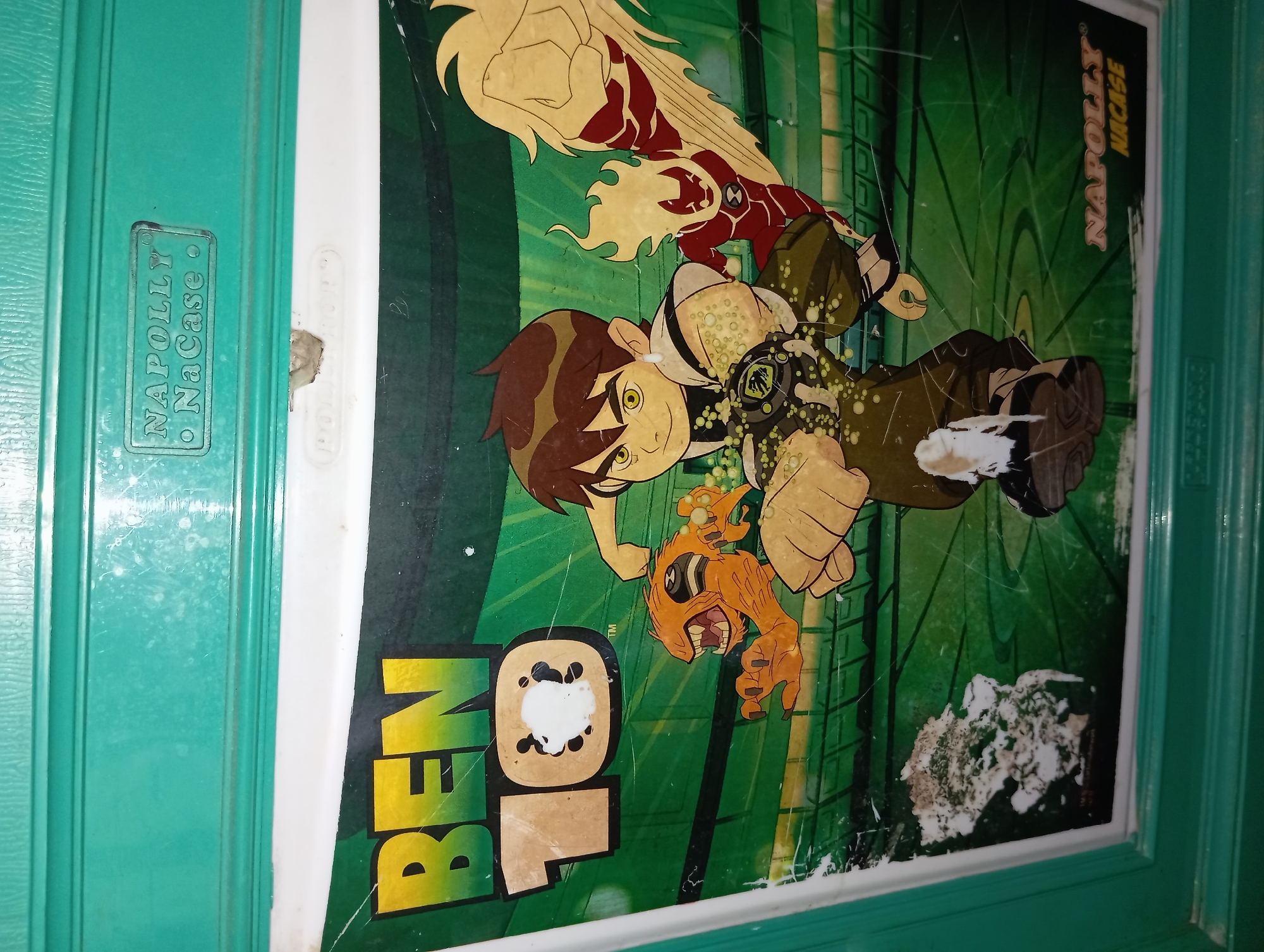 The image shows a close-up of a green plastic surface with an embedded graphic of the cartoon character Ben 10. The graphic features Ben Tennyson in his usual attire, along with two of his alien transformations: Heatblast and Four Arms. There are also two other alien characters present, one orange and one red with flames. The Ben 10 logo is prominently displayed. Text in a rectangular box at the top reads "NAPOLLY NaCase". Similar text appears at the bottom right corner. The plastic surface appears to be part of a container or a piece of furniture, with some scuff marks and peeling stickers visible. The overall scene suggests a manufactured item, likely a toy or storage box, featuring popular cartoon characters. This object is located in the city of Rejosari, Indonesia.