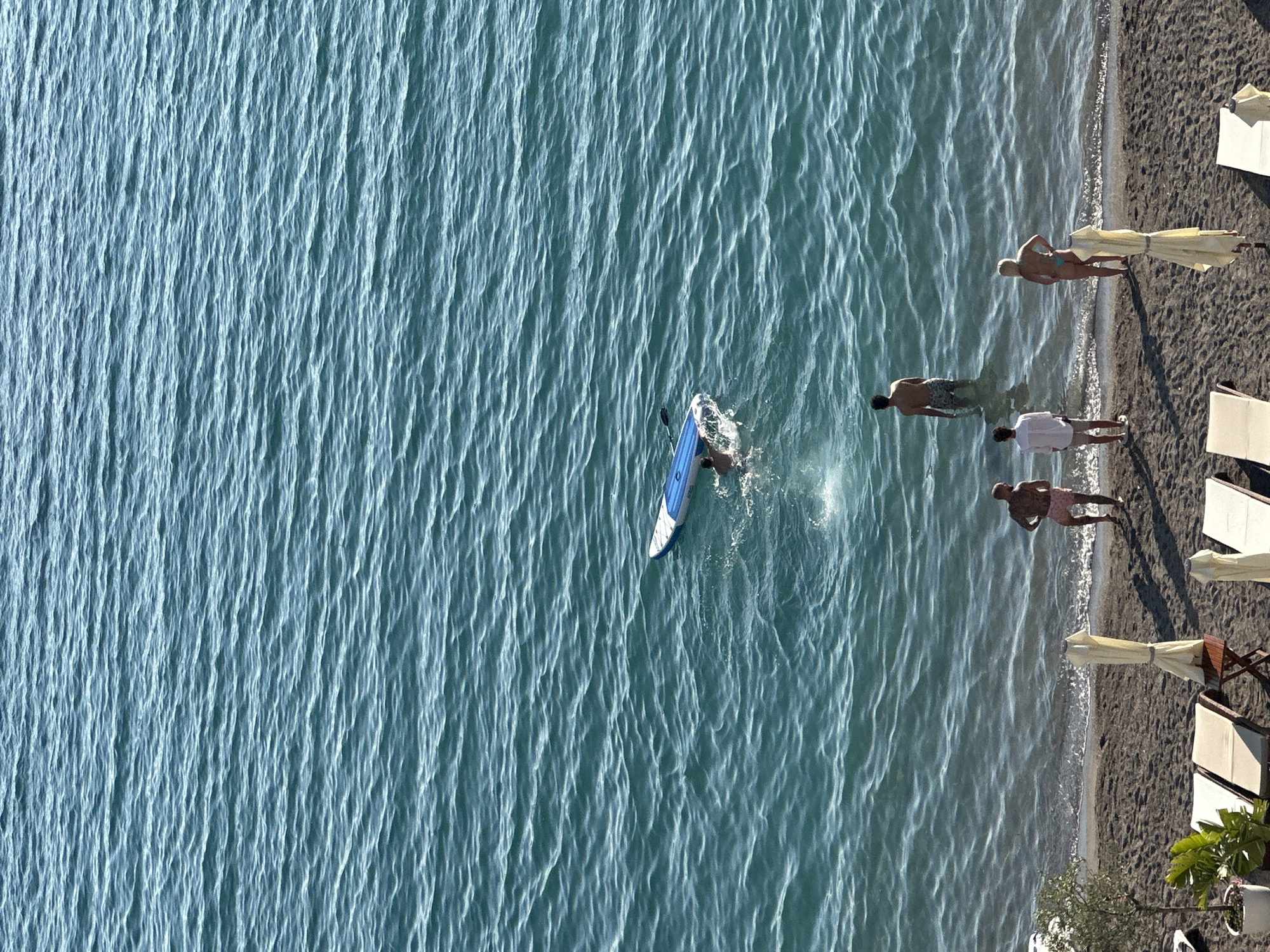 Aerial view of a beach in Budva, Montenegro. Several people are on the beach and in the water. One person is on a blue and white surfboard. The beach has umbrellas and chairs. The water is turquoise and has ripples.