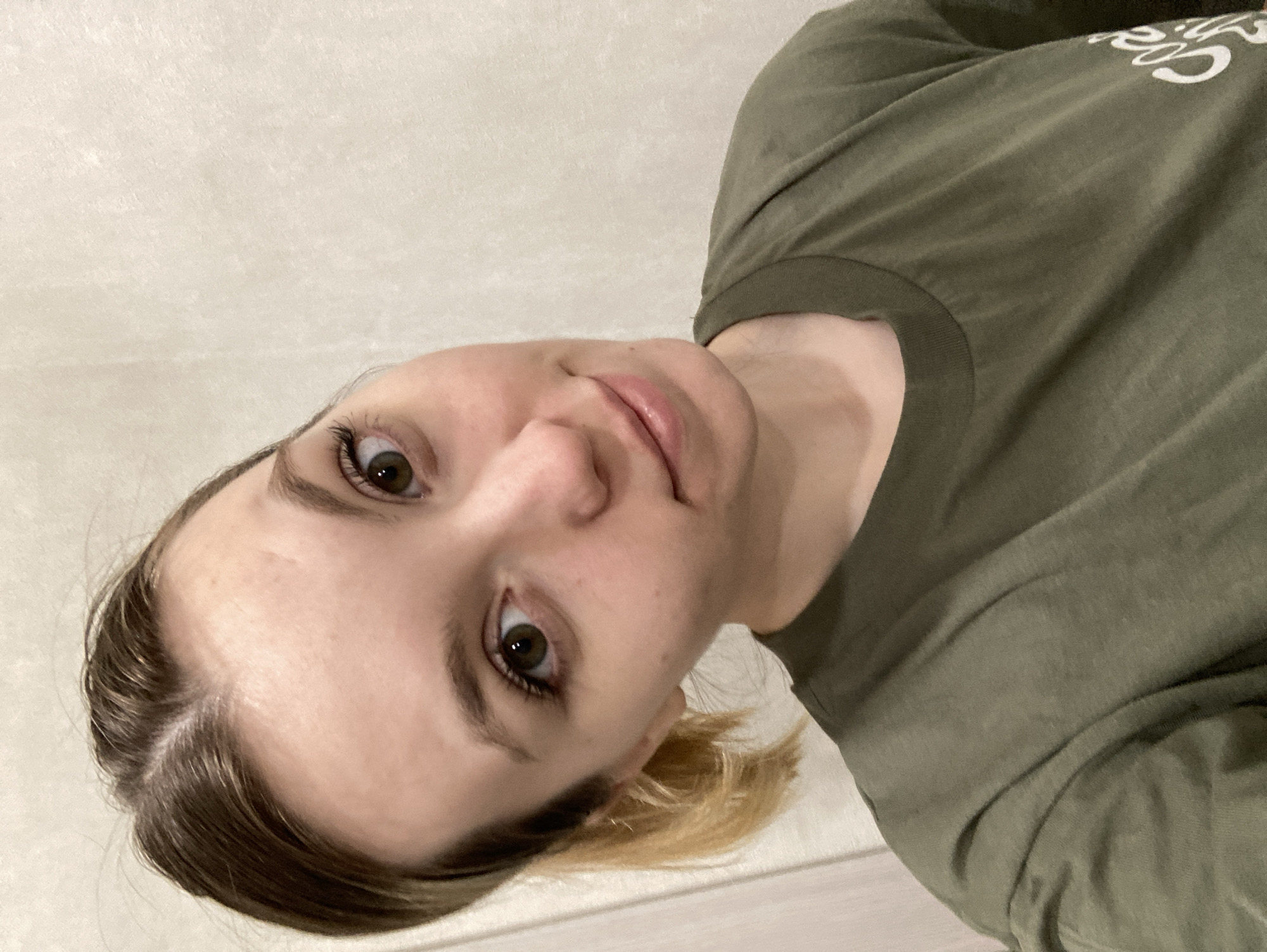 A selfie taken by a young woman. She is looking up at the camera with a slight smile. She has brown hair with blonde highlights. She is wearing a dark green t-shirt with white text on it. The background is a beige wall. This image was not necessarily taken in the city of Chaikovsky, Russia and I am unable to obtain such information.