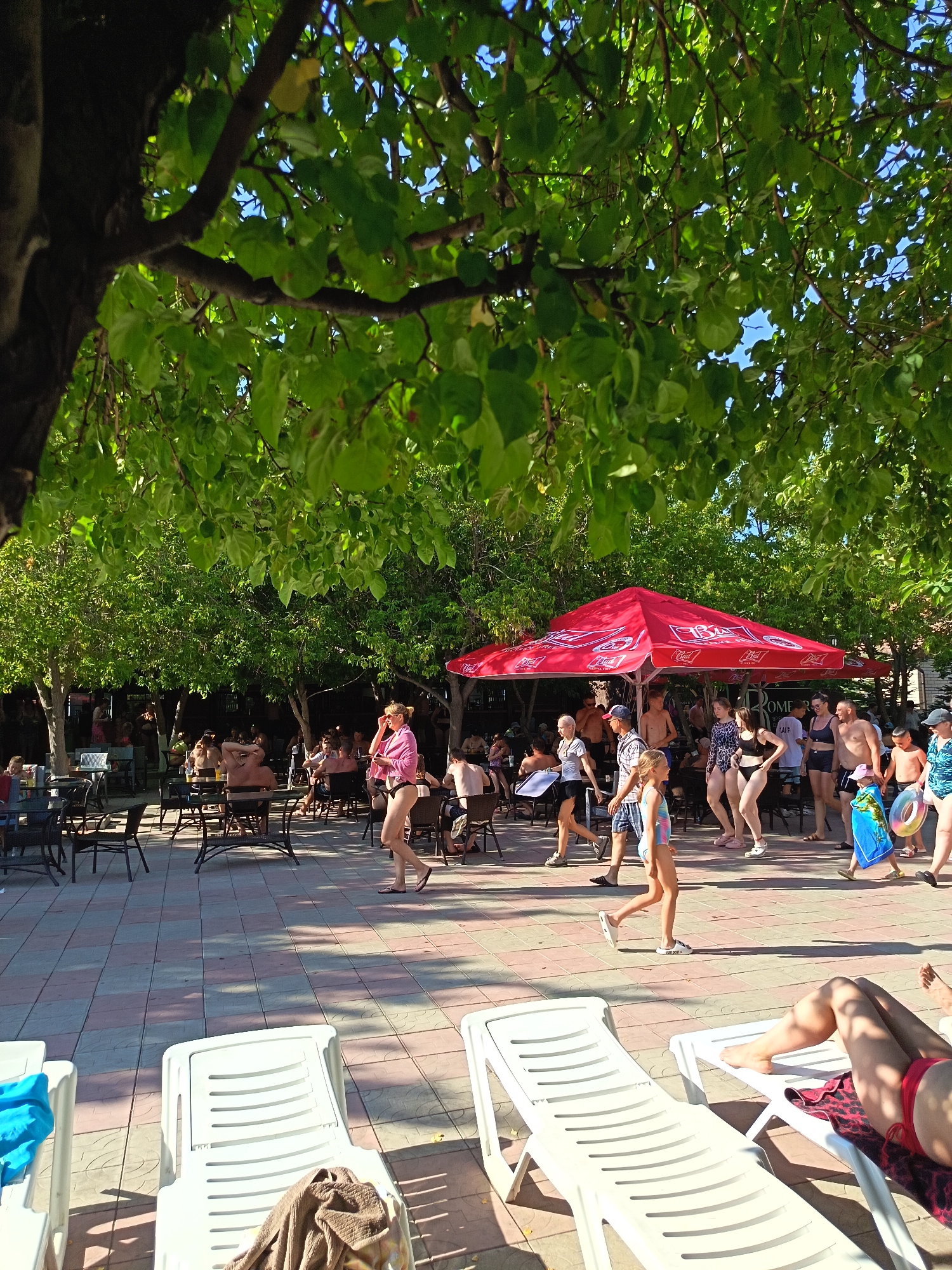 The image depicts a sunny outdoor scene at a beachside resort in Yarovoye, Russia, likely during the summer. In the foreground, two white plastic lounge chairs are visible on a tiled patio, one with a blue towel and the other with a brown towel. Beyond the chairs, a paved area with a patterned tile design separates the lounging area from a bustling section where people are walking and gathered.
A large red umbrella shades a seating area with tables and chairs, where numerous individuals are seated and conversing. Many people are dressed in swimwear, suggesting a recreational setting. To the right of the umbrella, a group of people are walking, some carrying beach accessories like a blue towel and a swim ring. The background is filled with trees, providing ample shade and contributing to the vacation atmosphere. The overall scene conveys a lively and relaxed resort environment.