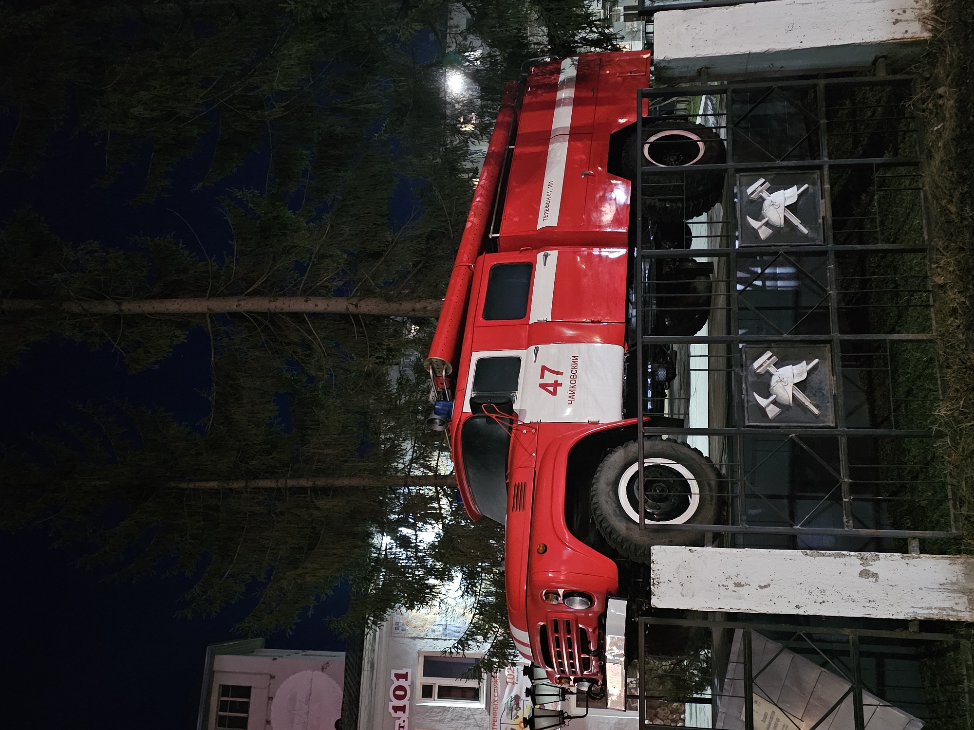The image displays a red and white fire engine, likely a historical model, parked outdoors at night. The vehicle is prominently featured, with its front facing towards the right side of the frame and its left side visible. It has a long ladder apparatus extending along its top.
Visible text on the side of the cab reads "47" and "ЧАЙКОВСКИЙ" (CHAYKOVSKIY), suggesting its affiliation with the city of Chaykovsky. On the rear side of the truck body, "ТЕЛЕФОН 01, 101" (TELEPHONE 01, 101) is written, indicating emergency phone numbers.
The fire engine is positioned next to a black metal fence or gate. Integrated into the fence are several square panels, each containing a relief of a fire axe and another tool, possibly a helmet or nozzle, emphasizing the firefighting theme. Beyond the fence, a dark background of evergreen trees suggests a park or forested area.
In the background, on the far left, a light-colored building is visible. A sign on this building shows "Т.101" (T.101 or Tel. 101), reinforcing the emergency services context. Another partially obscured sign on the building has text beginning with "ВЕННЫХ СЛУЖБ...", which is likely part of "ЭКСТРЕННЫХ СЛУЖБ" (EMERGENCY SERVICES).
The scene is illuminated by artificial light sources, casting a glow on the fire truck and parts of the fence, contrasting with the dark night sky and the shadowy trees. There are no people visible, and the overall impression is that of a static display or monument to firefighting services.