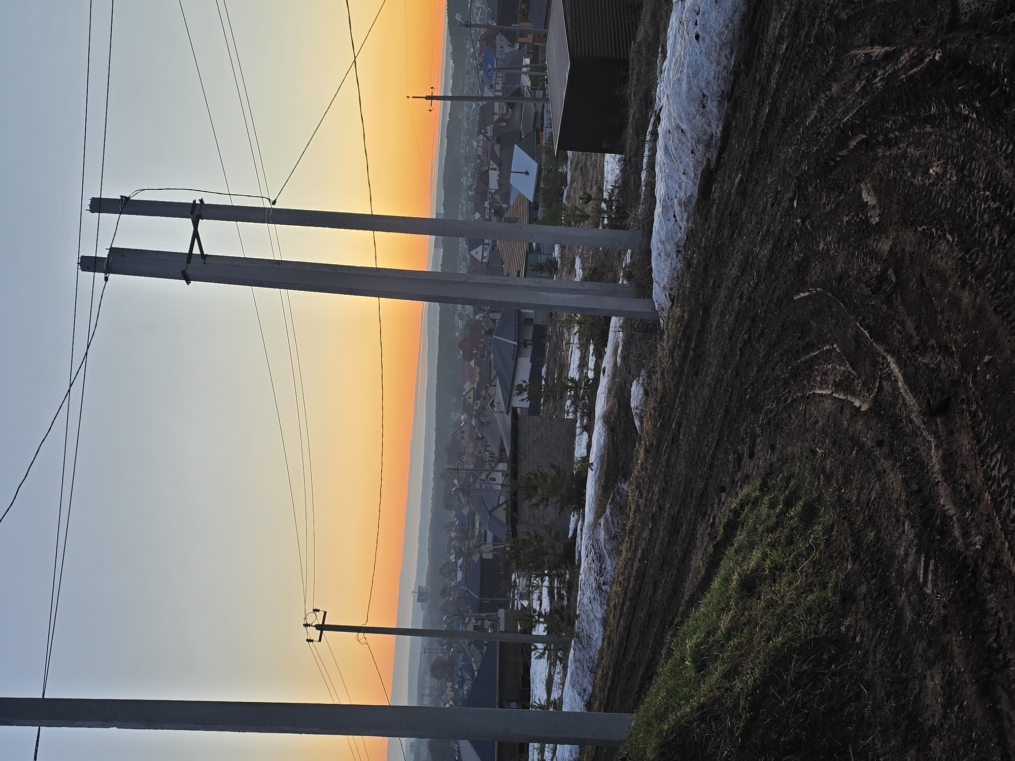 The image shows a view of a residential area during what appears to be either a sunrise or sunset, characterized by an orange glow in the sky. The location is Chaikovsky, Russia, as per the prompt. In the foreground are concrete utility poles carrying electrical wires, typical of rural or suburban landscapes. The ground in the foreground is a mix of dirt and patchy green grass, with some remaining snow in shadowed areas, indicating a transitional season like late winter or early spring. Beyond the utility poles, a dense collection of houses with varying roof colors and architectural styles can be seen, suggesting a mixture of older and newer constructions. The overall environment has a hazy, slightly misty quality. There are no people visible in the scene.