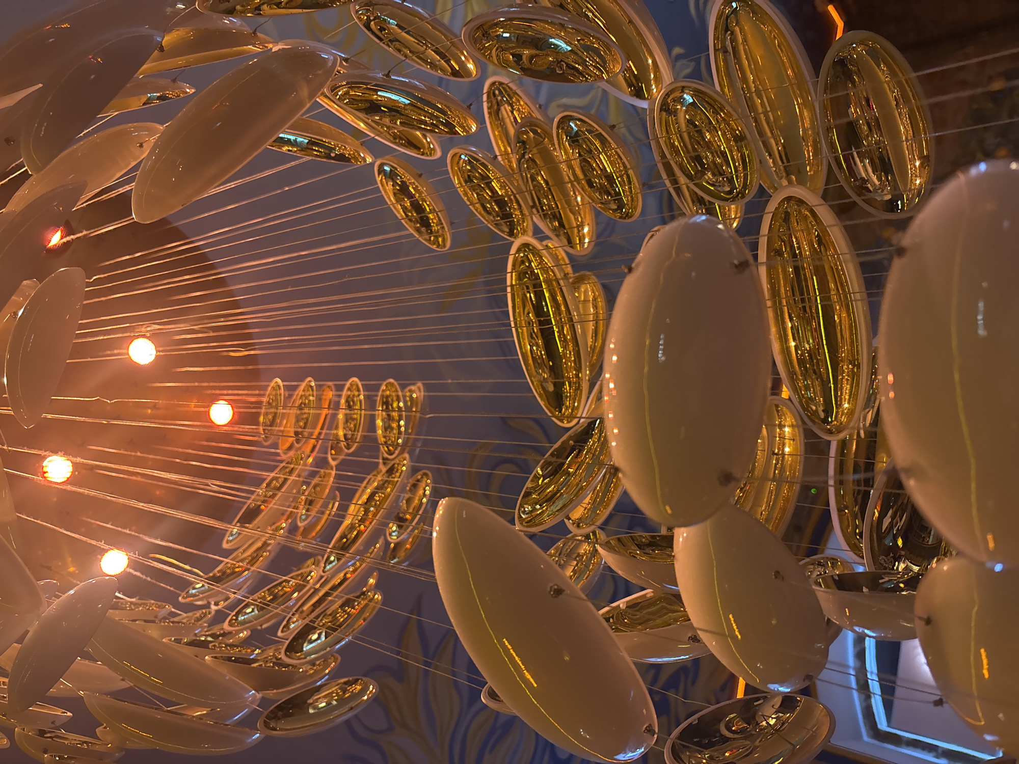 This is a low-angle shot of a contemporary chandelier with multiple oval-shaped elements. The chandelier is illuminated by warm, orange lights, which create reflections on the glossy surfaces of the hanging elements. Some elements are white and opaque, while others are gold and highly reflective. The background shows a ceiling with a blue and gold patterned mural. The scene is indoors and appears to be a decorative element in a public space or an establishment. No people are visible in the image. The location is not identifiable from the image content.