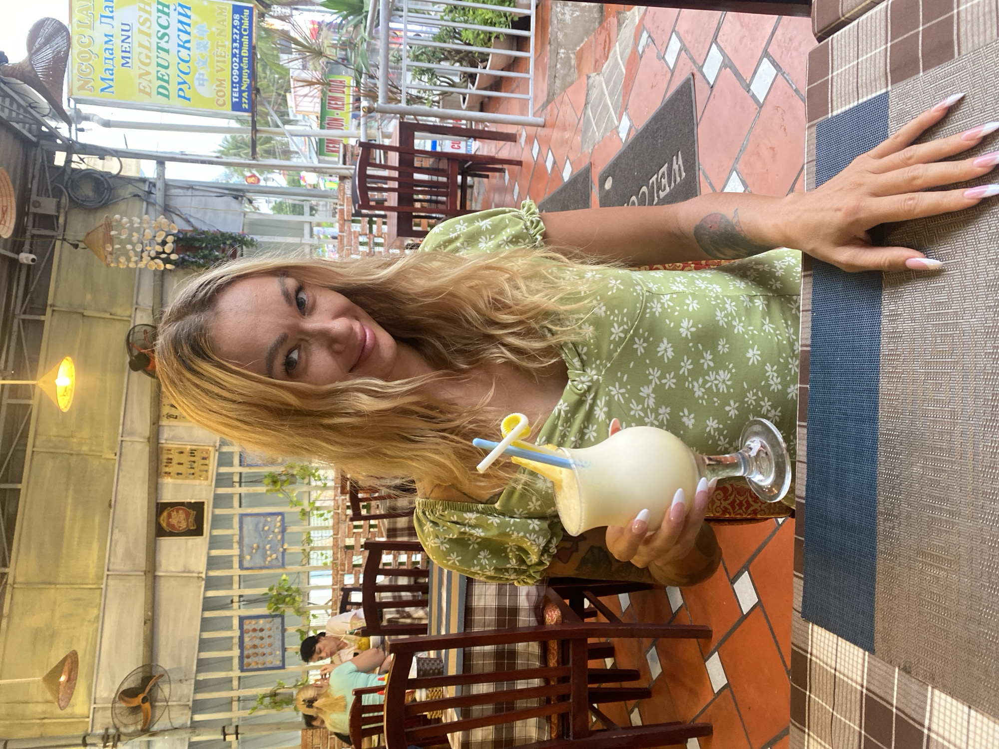 A woman in a green floral dress is seated at an outdoor table in Phu Thuy Ward, Vietnam, holding a white cocktail in a glass. Her blonde hair is partially framing her face as she smiles. Her left arm is extended towards the right side of the image with her hand resting on the table. The setting appears to be a restaurant or cafe, as indicated by the tables and chairs. A sign in the background shows a menu in various languages, including English, German, and Russian, as well as Vietnamese. The address is "27A Nguyễn Đình Chiểu" indicating the location of the establishment. Other people are visible in the background.