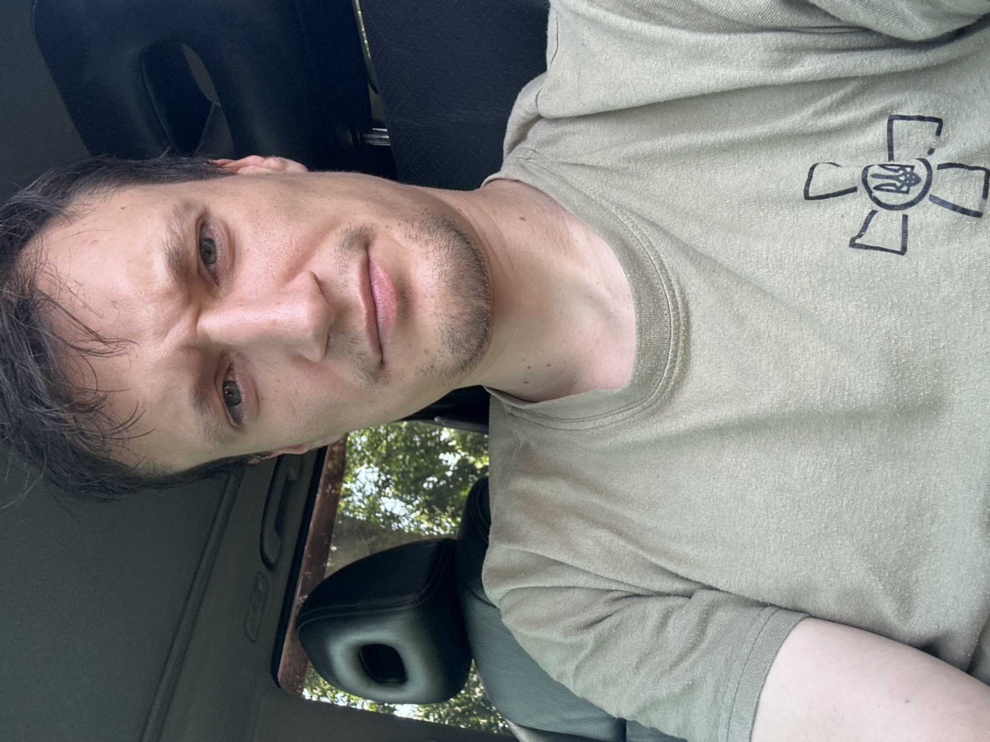 A close-up, high-angle, selfie-style photograph shows a man from the chest up, seated in the driver's seat of a vehicle. The man has short, dark hair, light skin, and a beard stubble. He is wearing a tan t-shirt with a black emblem of a cross with a trident in the center, a symbol associated with Ukraine. The interior of the vehicle, including a black leather headrest and a portion of the dashboard, is visible. The background shows greenery, suggesting the vehicle is outdoors, possibly near Makiv, Ukraine. The man's facial expression is neutral, and he is looking directly at the camera. The lighting is natural and appears to be daylight.