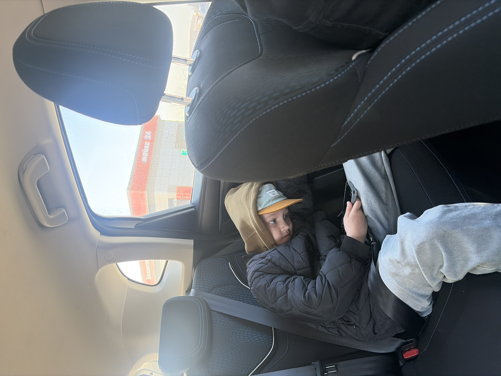 A young individual, appearing to be male, is seated in the rear passenger-side seat of a vehicle in Saint Petersburg, Russia. The individual wears a black quilted jacket, light blue jeans, a tan hooded sweatshirt, and a light blue baseball cap with a beige brim. They are secured by a black seatbelt with a red buckle release and are holding a dark-colored smartphone in both hands. The individual's head is tilted and resting on their left arm, looking towards the camera. The vehicle's interior features dark fabric seats with light blue stitching, and a light-colored ceiling with a grab handle and an "AIRBAG" label visible on the B-pillar. Through the side windows, an outdoor commercial scene under bright daylight is visible. Two adjacent buildings with red roofs and red trim are present. The building on the left has partially visible white Cyrillic text "Бесконто," indicating a contactless service. The building on the right prominently displays "Мойка 24" (Car Wash 24) in white Cyrillic lettering, identifying the location as a 24-hour car wash facility.