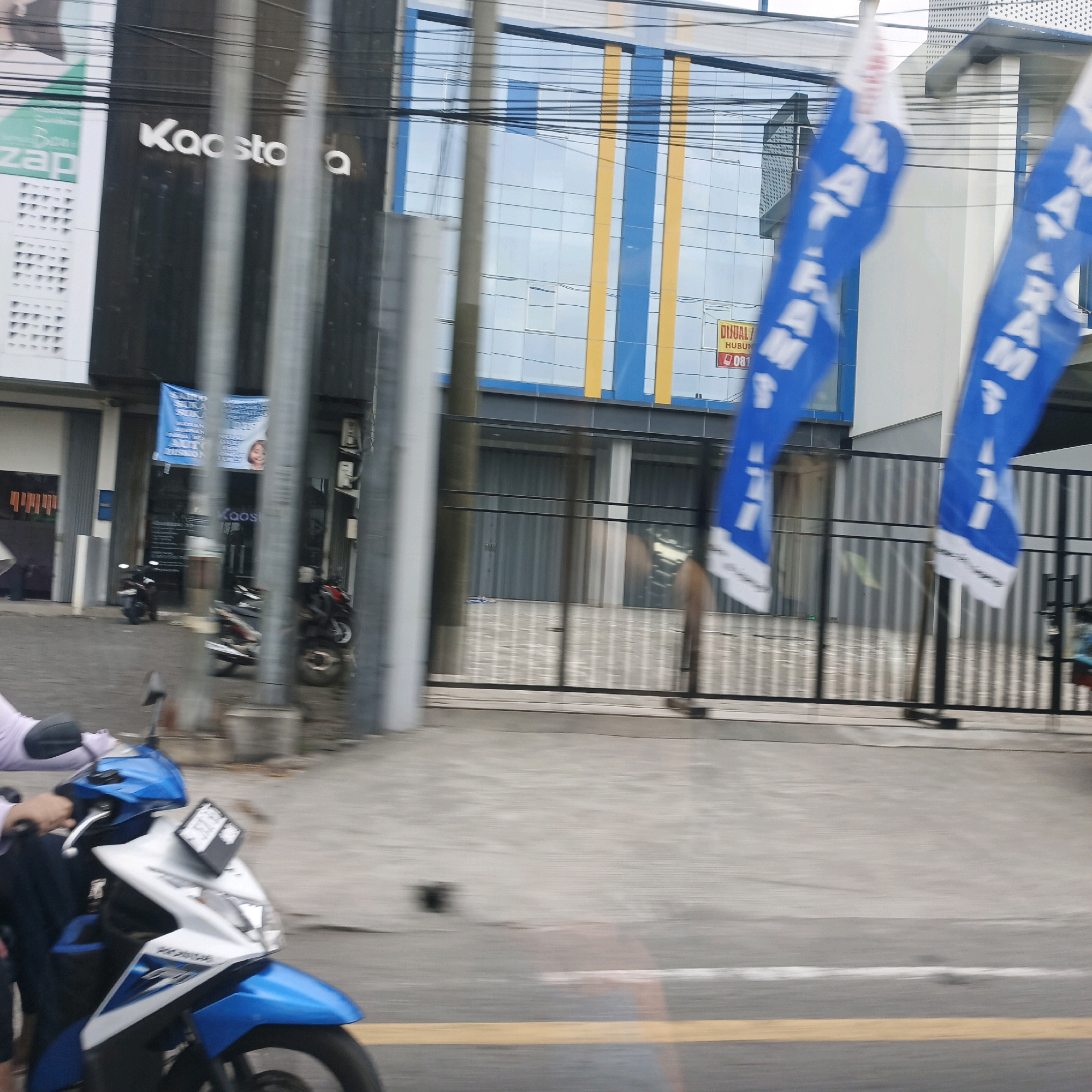The image captures a street scene in Semarang, Indonesia, likely taken from a moving vehicle. In the foreground, a blue and white motorcycle is partially visible, with a person's arm and hand on the handlebars, indicating they are riding. Two blue flags with white lettering are prominently displayed, fluttering in the wind. Behind these flags, a building with a modern glass facade featuring blue and yellow vertical accents is visible. Signs are present on the building, including one that reads "KAOSTO A". To the left, another building section with a dark facade and the word "zap" is seen. Various poles and wires crisscross the upper portion of the image, suggesting an urban environment. The ground appears to be paved, with a yellow line marking a road. Several motorcycles are parked or in motion in the background, alongside a closed storefront. The overall impression is a snapshot of everyday life in a commercial area of Semarang.
