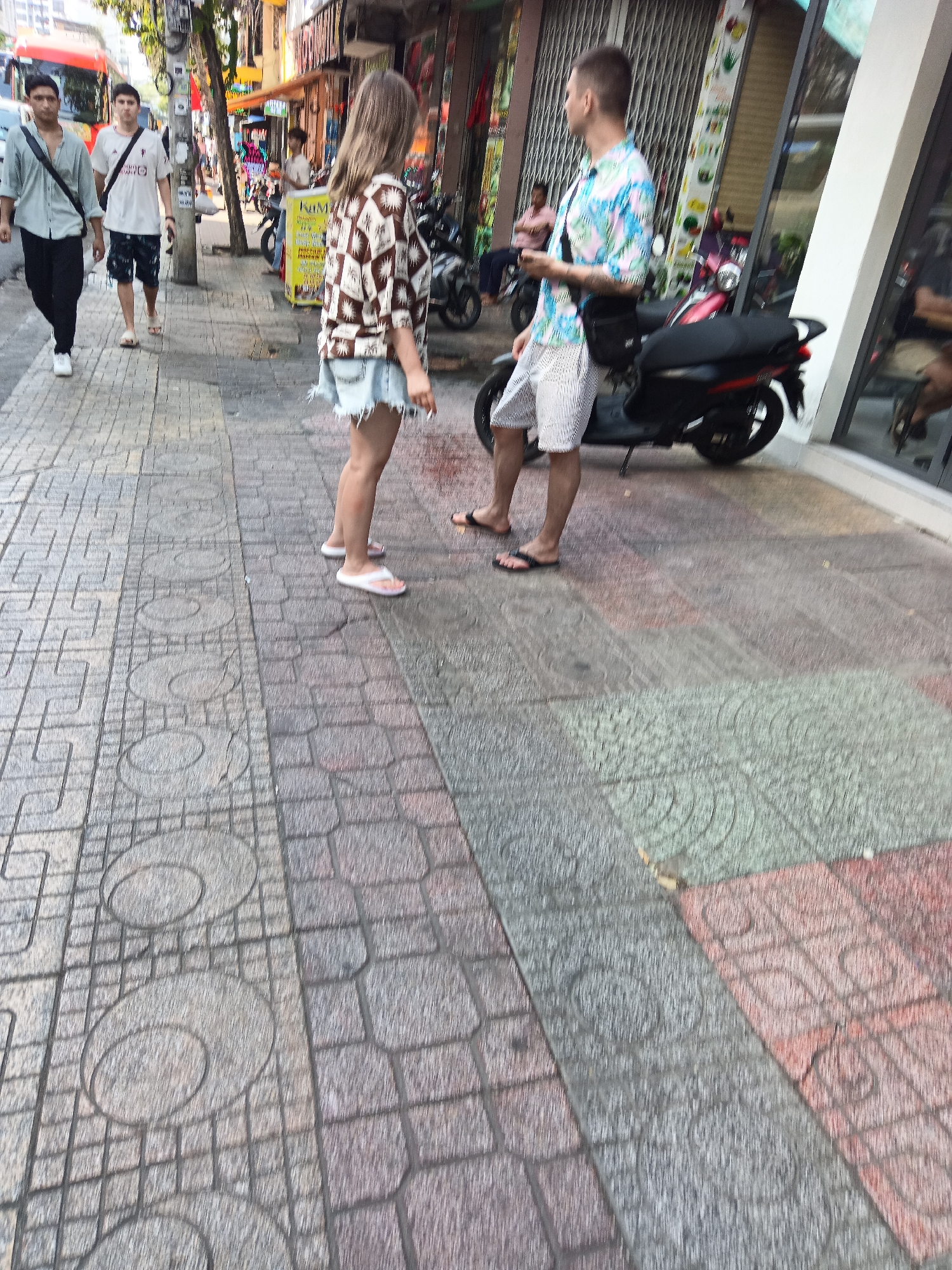 Two young adults are standing on a sidewalk in Nha Trang, Vietnam, facing each other and conversing. The male, on the right, wears a colorful, short-sleeved patterned shirt and striped shorts, with a black messenger bag slung across his chest. The female, on the left, has long blonde hair and is dressed in a brown and white patterned jacket over a denim skirt and white flip-flops. She is holding a white object, possibly a cigarette, in her right hand.
Behind them, a black scooter is parked, and further in the background, shops with open doorways and display windows line the street. Another young man, wearing a light blue long-sleeved shirt and black pants, walks away from the camera on the left side of the sidewalk, with a dark bag over his shoulder. A second young man in a white t-shirt and black shorts walks beside him. The sidewalk is paved with textured tiles. The overall scene suggests a casual daytime interaction on a bustling city street.