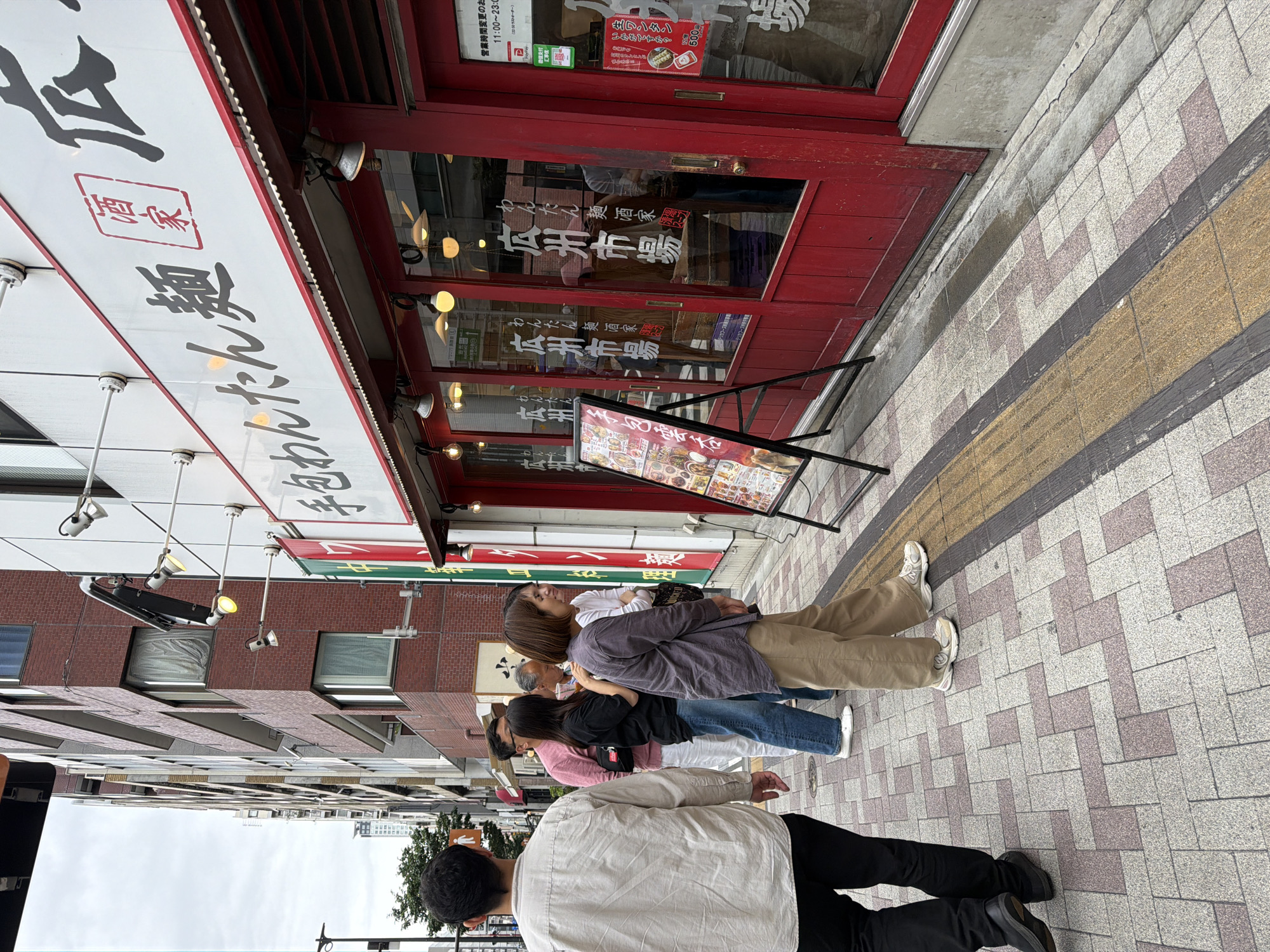 A group of people stand outside a restaurant with red doors on a cloudy day in Meguro, Japan. The restaurant has a white sign with Japanese writing and red accents. A menu board is propped up in front of the restaurant. The sidewalk is paved with patterned tiles. The people in the image are dressed casually. One man in the foreground is walking away from the camera, wearing a light-colored button-down shirt and black pants. Behind him, a woman in a grey shirt and tan pants is looking towards the restaurant. A man in a pink shirt and white pants, and a woman in a black t-shirt and blue jeans stand next to her. Another person, partially obscured, stands behind them. The street appears to be a pedestrian walkway alongside a building.