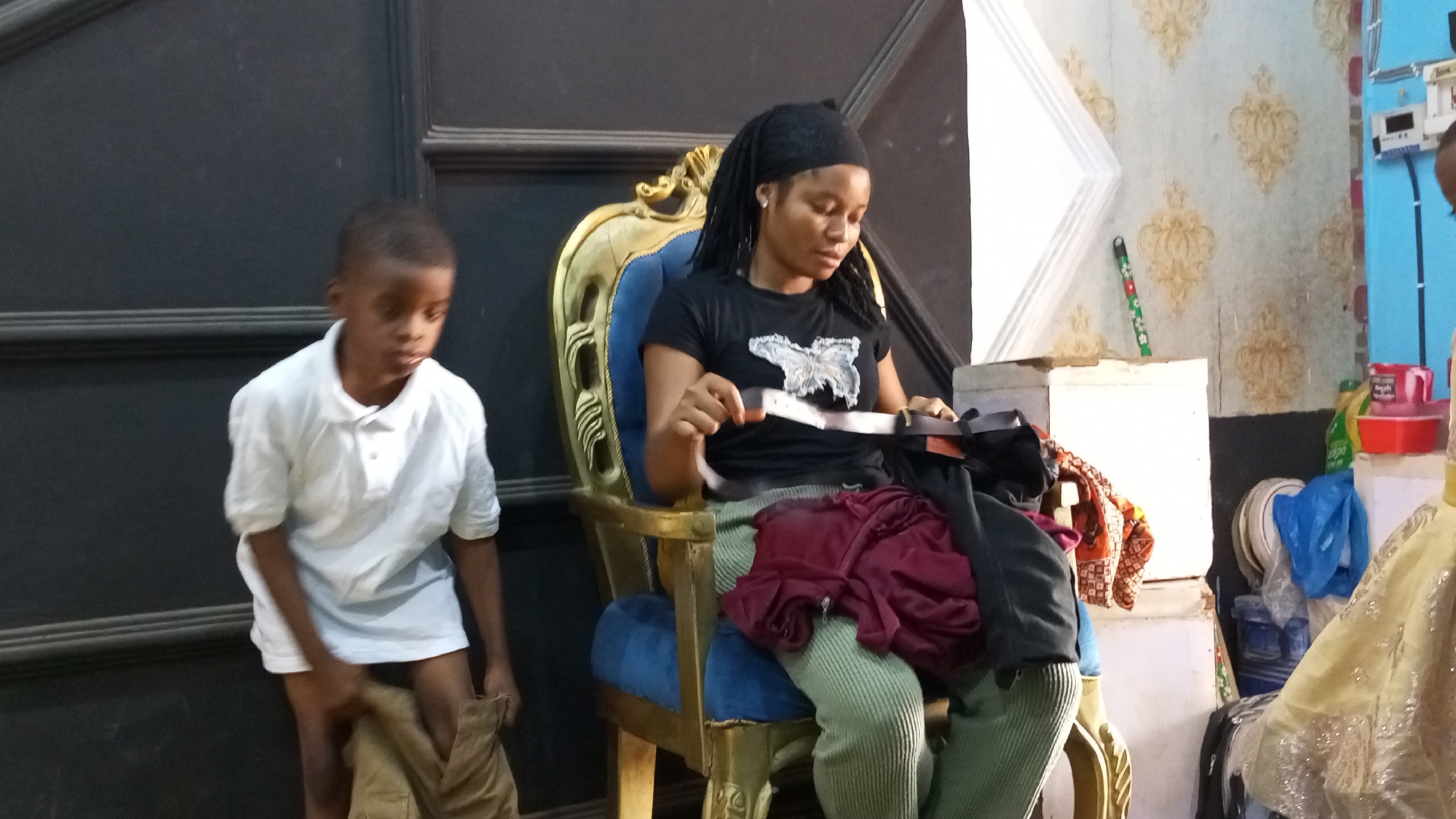 In Sabon Tasha, Nigeria, a young boy stands near a dark wall, pulling on khaki-colored trousers. He wears a white polo shirt. To his right, a woman sits in a blue and gold ornate chair. She wears a black t-shirt, green pants, and a black head covering. She is holding a white belt. In front of her are red and black pieces of clothing. Behind her is a room with patterned wallpaper and household items.