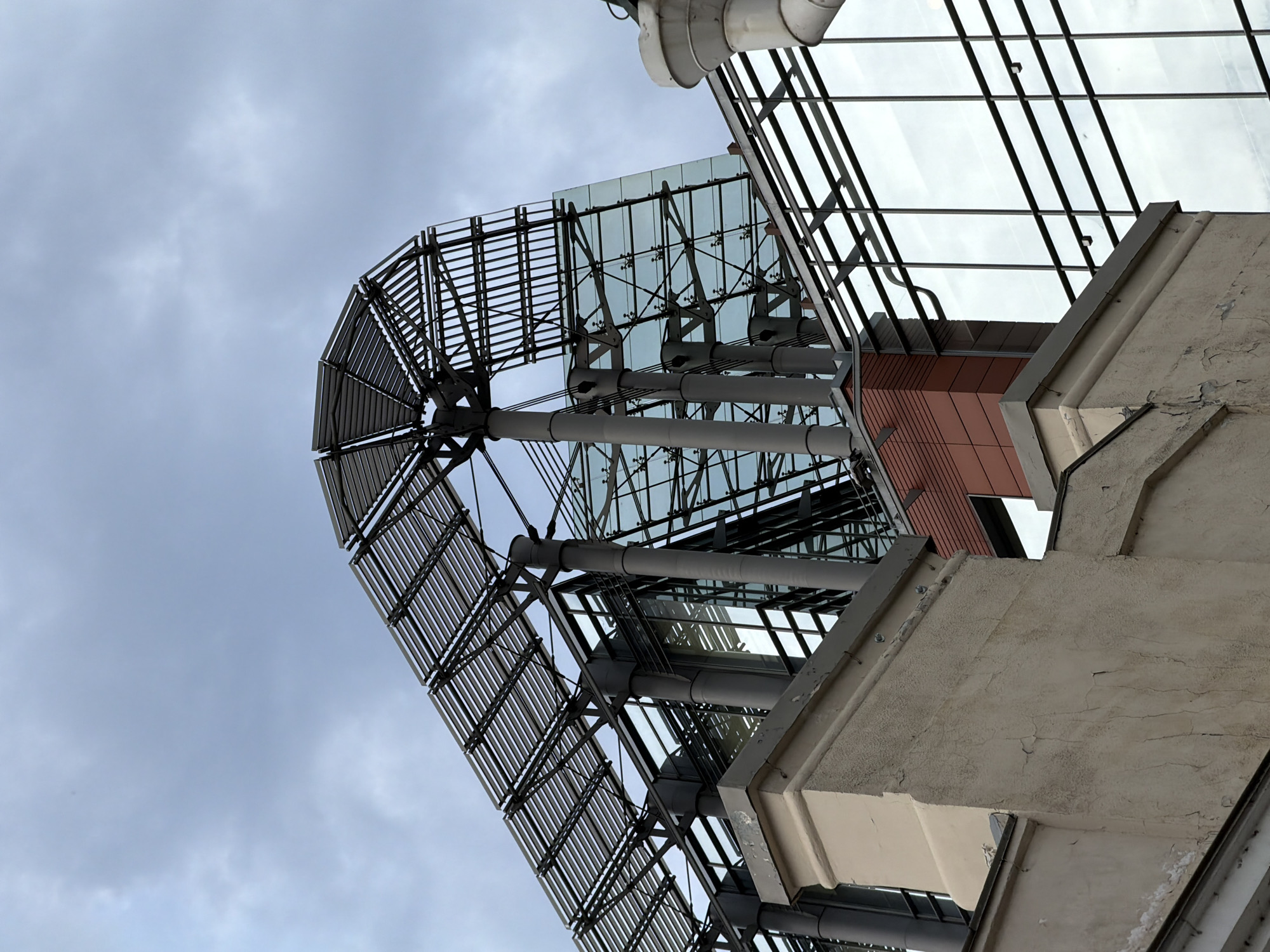 A low-angle shot reveals the architecture of a building, blending metal, glass, and weathered concrete elements under a cloudy sky. The composition is dominated by the upward view, obscuring any specific location information. A metal structure with a curved, louvered facade arcs out from the building's base, contrasting with the geometric lines of the glass and metal framework above. The building's exterior shows signs of age, with peeling paint and visible cracks in the concrete.
