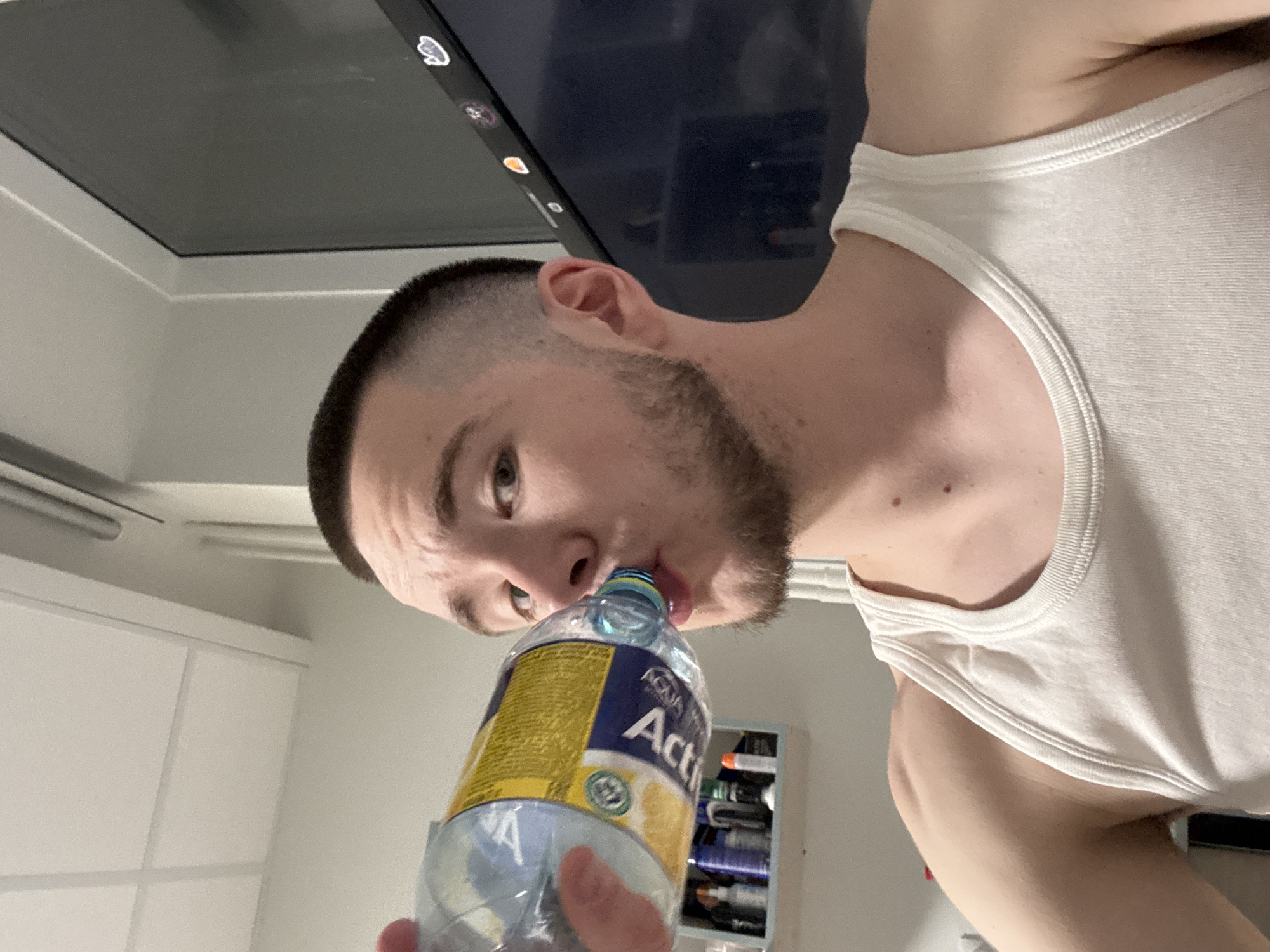 A close-up image captures a young man drinking from a plastic water bottle, viewed from a low angle. The man has short, dark hair styled in a fade or buzz cut, and a visible beard or stubble. He is wearing a white sleeveless tank top. His eyes are directed towards the camera as he brings the bottle to his lips.
The water bottle, held in his hand, has a prominent blue and yellow label on which the text "AQUA Acti" is clearly visible, along with a small graphic of a lemon. The background consists of a dark monitor or screen with a few white icons along its top edge, suggesting a desk or computer setup. Behind him, white walls and a ceiling are visible, along with a small shelving unit containing other bottles or items on the right side of the frame. The overall setting appears to be an indoor room with ambient lighting. The image is notably rotated, with the subject appearing sideways in the frame.
The man's expression is neutral, focused on drinking. No specific time of day or weather conditions can be discerned from the indoor setting. The location context provided is Kazan, Russia.
