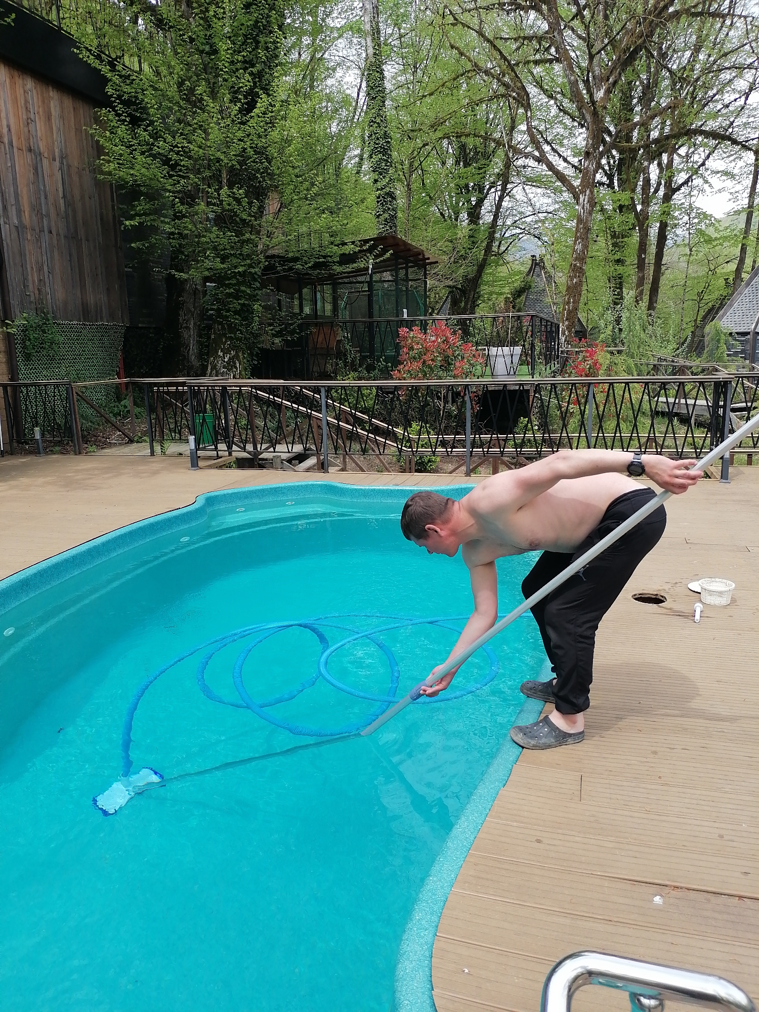 A shirtless man, wearing black pants and dark clogs, is captured in an outdoor setting, actively cleaning a swimming pool. He is bent over the edge of the pool, holding a long white pole with a cleaning head submerged in the teal-blue water. A blue hose, partially coiled in the pool, connects to the cleaning apparatus. A dark watch is visible on his left wrist.
The swimming pool has an irregular shape and is surrounded by a light brown wooden or composite deck. On the deck, near the man, a white skimmer basket, a white pipe fitting, and a dark circular opening are visible. Part of a silver metal handrail, likely for pool access, can be seen in the bottom right corner of the frame.
Beyond the pool deck and a black metal railing with a decorative criss-cross pattern, a lush natural environment unfolds. Dense green trees with new spring foliage dominate the background, some revealing their trunks and branches. Red flowering bushes add a touch of color behind the railing. Rustic wooden structures, possibly cabins or outbuildings with dark roofs, are nestled among the trees, suggesting a natural or resort-like setting. A tall wooden wall is visible on the far left.
The scene is illuminated by bright, diffused daylight, indicating an overcast or partly cloudy sky. The man appears focused on his task of pool maintenance. The overall ambiance suggests a peaceful, natural location, consistent with the provided context of Medoveevka, Russia. There is no discernible text in the image.