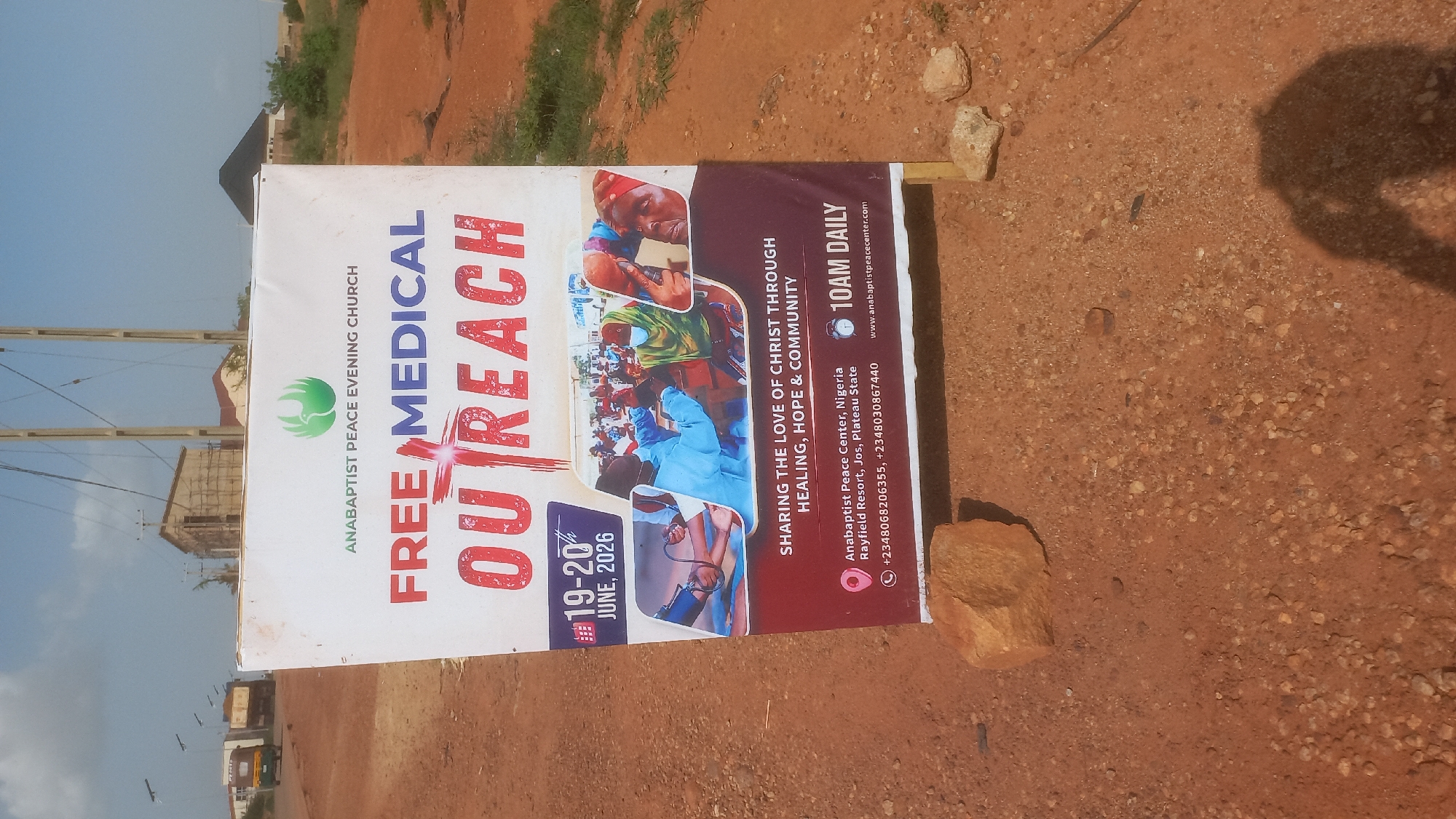 This is a vertical, outdoor photograph of a banner advertising a "Free Medical Outreach" organized by the Anabaptist Peace Evening Church. The banner is the central subject, set against a backdrop of a dusty, reddish-brown ground with some scattered stones and pebbles. In the background, a few buildings and trees are visible, along with power lines. The sky appears bright, suggesting daylight, and the overall impression is of a somewhat arid or rural environment.
The banner itself features bold text, with "FREE MEDICAL" prominently displayed in red and blue, followed by "OUTREACH" in large, red, stylized lettering with a cross symbol integrated into the word. Below the main text, dates "19-20 JUNE, 2026" are shown, along with a time indication of "10AM DAILY."
The banner also includes several images of people, depicting medical professionals attending to individuals, suggesting the nature of the outreach. Text provides further details about the event's purpose: "SHARING THE LOVE OF CHRIST THROUGH HEALING, HOPE & COMMUNITY." Contact information for the "Anabaptist Peace Center, Nigeria," located at "Rayfield Resort, Jos, Plateau State," is also present, along with phone numbers and a website address. The presence of the banner, likely set up for public viewing, indicates an upcoming or ongoing event intended to offer medical services and community support.