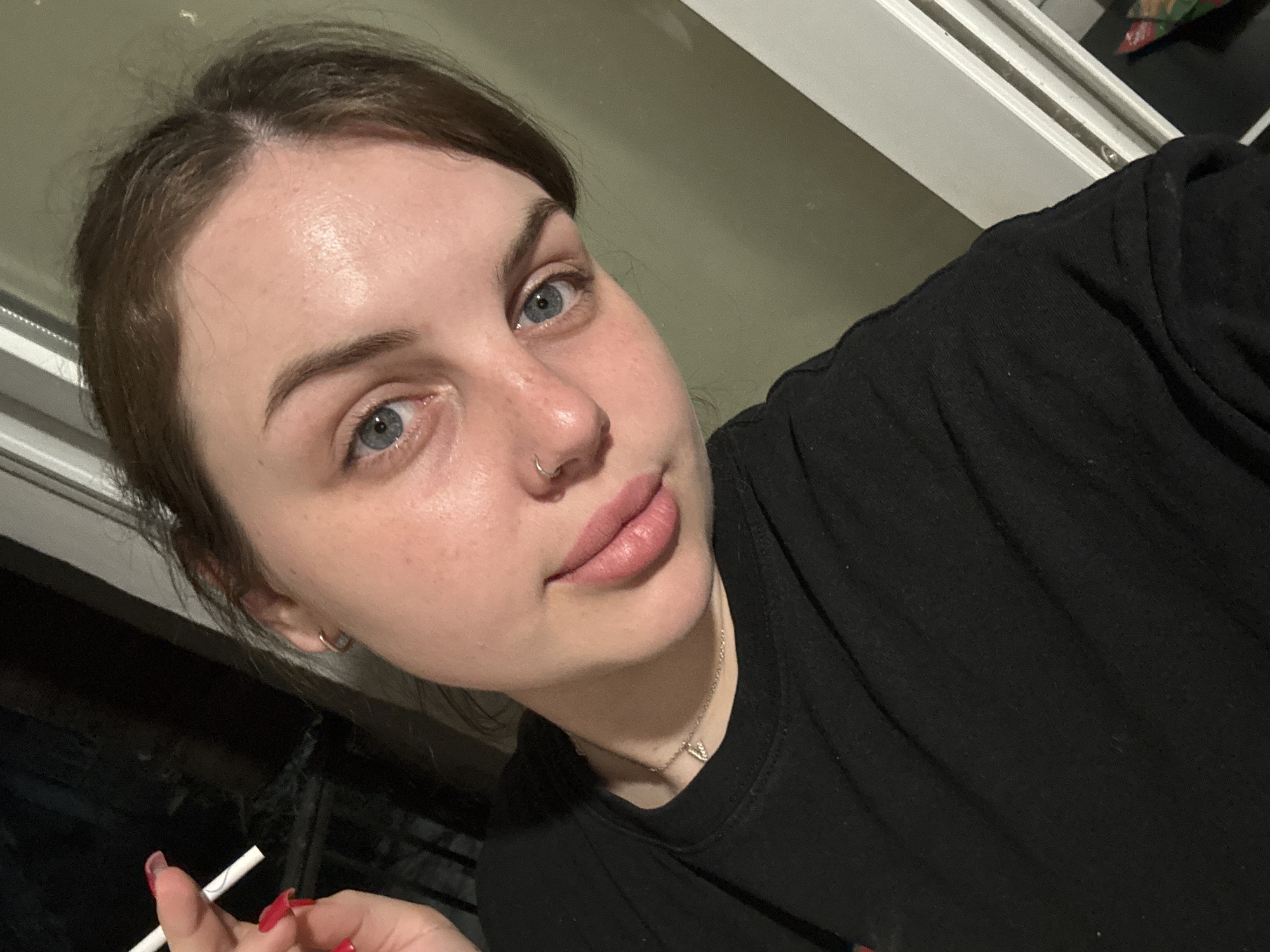 A close-up, high-angle shot shows a fair-skinned woman with brown hair and blue eyes, wearing a black shirt and a silver necklace. She has a nose ring in her left nostril, small hoop earrings, and red nail polish. She is holding a thin white object, possibly a cigarette or straw, between her fingers. The background includes a window frame and a dark, indistinct area. No information in the image indicates that this photo was taken in Chornomorskoe, Ukraine.
