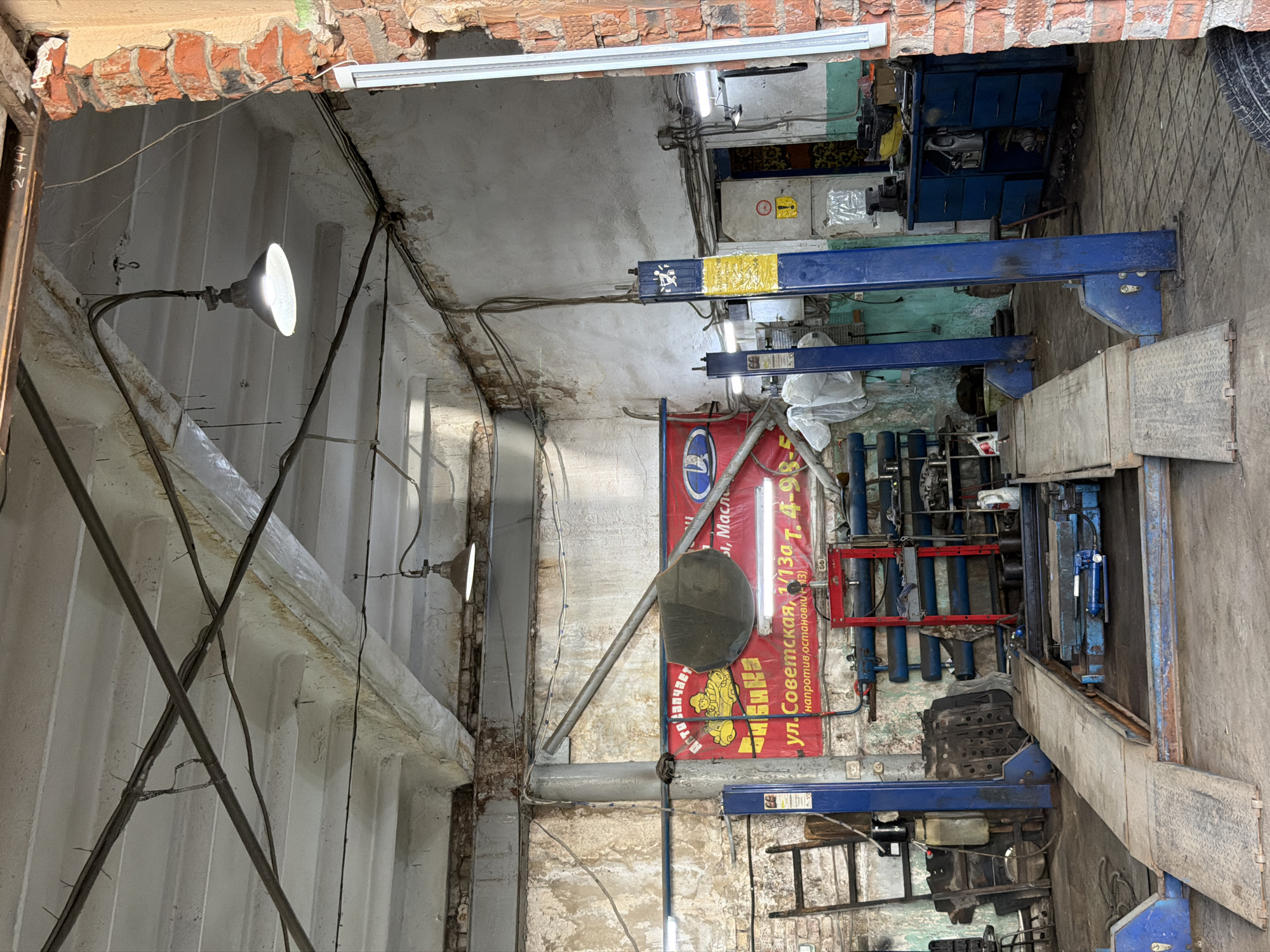 The image shows the interior of a repair shop in Chaikovsky, Russia. The interior is a garage. It has two car lifts and a sign that says "ул. Советская, 1/130 т. 4-98-5 напротив остановки + 13". There is a bare lamp in the top left. The interior is a state of disrepair.