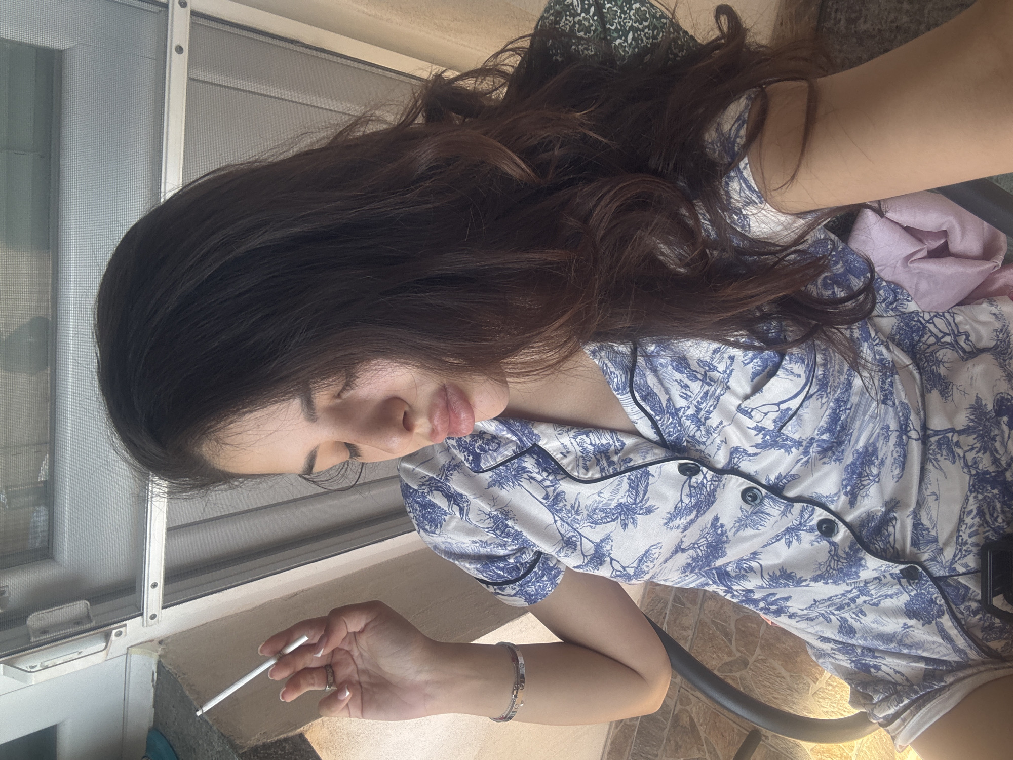 The image shows a young woman with long, dark, wavy hair, sitting in a chair outdoors. She is wearing a blue and white patterned pajama set. Her eyes are closed, and her lips are slightly parted as if she is exhaling smoke. She is holding a cigarette between her index and middle fingers. The background shows a screen door, and a tiled floor. There is no indication of the specific location within the city of Plovdiv, Bulgaria.