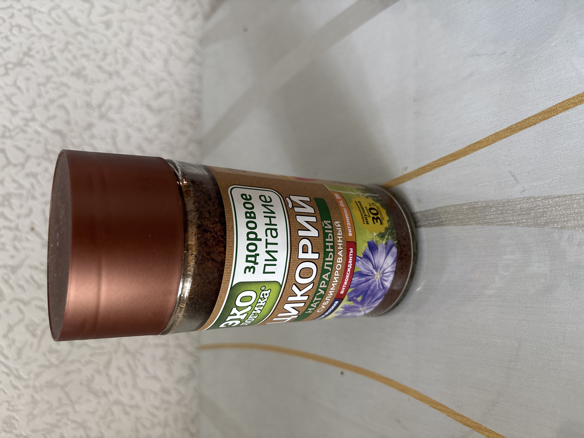 A close-up, high-angle shot shows a glass jar of chicory on a patterned surface. The jar has a metallic copper lid and a label with green and white text. The label features the words "ЭКО ЛОГИКА" (Eco Logic) and "ЗДОРОВОЕ ПИТАНИЕ" (Healthy Nutrition). Below this, "ЦИКОРИЙ" (Chicory) is prominently displayed, followed by "НАТУРАЛЬНЫЙ СУБЛИМИРОВАННЫЙ" (Natural Sublimated). The label also indicates the presence of antioxidants and vitamins B6, B9, and states "СОДЕРЖАНИЕ ИНУЛИНА более 30%" (Inulin content over 30%). A stylized drawing of a blue chicory flower is visible on the label. The chicory powder inside the jar is a dark brown color. The surface beneath the jar appears to be a table with a textured pattern of light and dark lines, with two prominent yellow-gold stripes running diagonally. The background is a textured, off-white wall. No people are present. The scene is indoors.