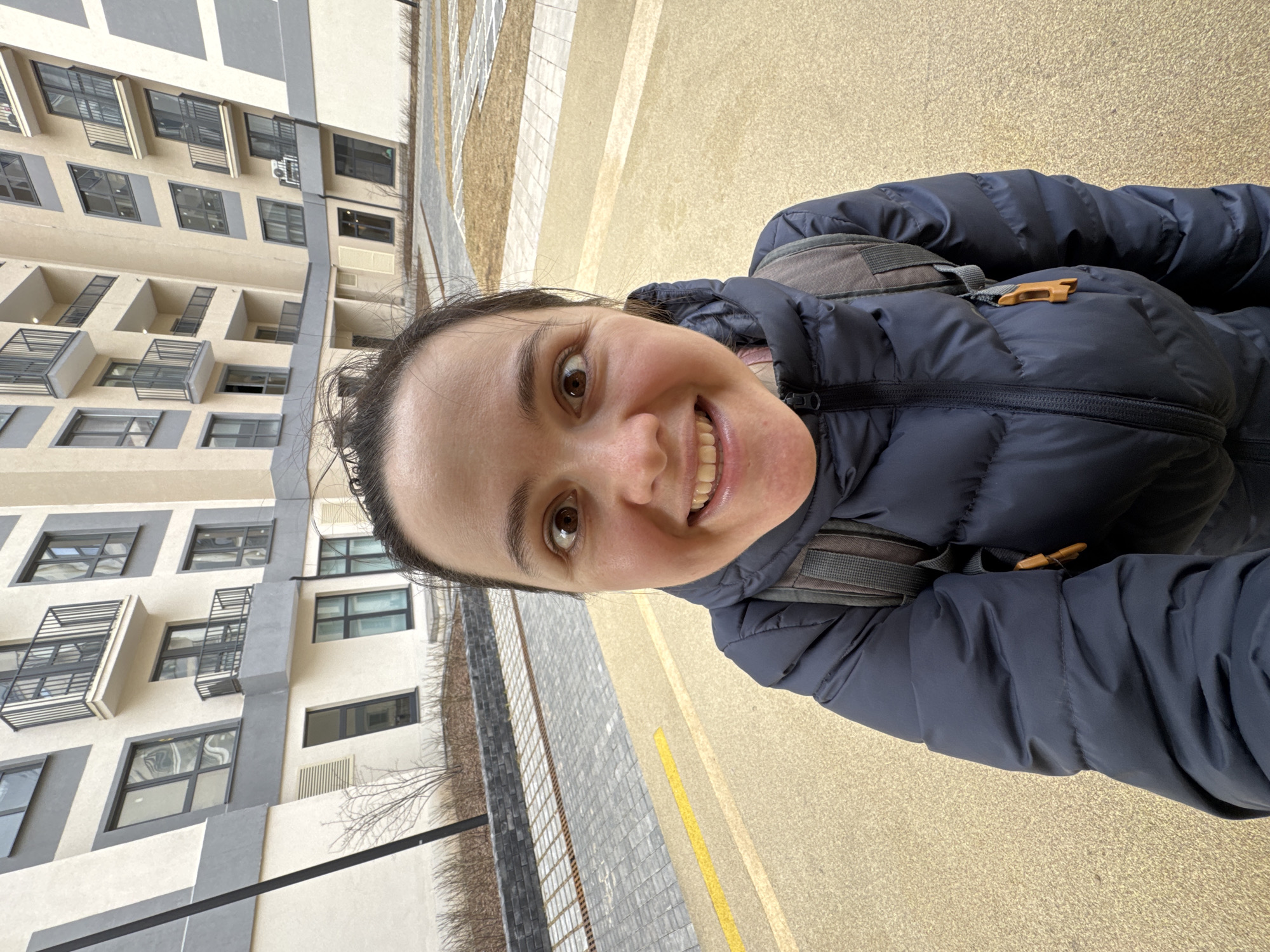 A woman in a navy blue puffer jacket and a gray backpack is taking a selfie outdoors. She is smiling at the camera. Behind her is a light beige building with gray accents and black framed windows. A gray stone wall is next to a yellow pavement. The location is the city of Izhevsk, Russia.
