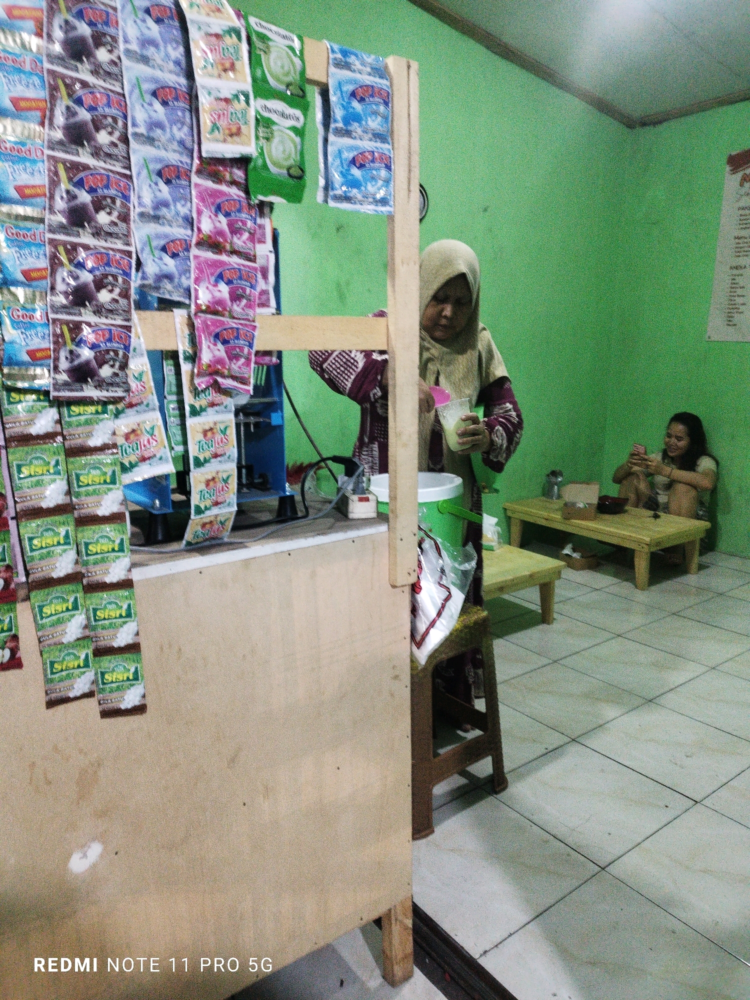 The image depicts an indoor scene with a green-painted wall and a tiled floor. In the foreground, a wooden counter displays numerous small, colorful packets of various food or drink products hanging from wooden pegs. A woman wearing a hijab is standing behind the counter, holding a plastic cup with a pink lid. To the right, a younger woman sits on a low wooden table, looking at her pink smartphone. Another low wooden table is visible in front of her. The environment appears to be a small shop or food stall. The text "REDMI NOTE 11 PRO 5G" is visible in the bottom left corner, indicating the device used for capture. While the location is identified as Saruni, Indonesia, no specific landmarks or context within the city are visually present.