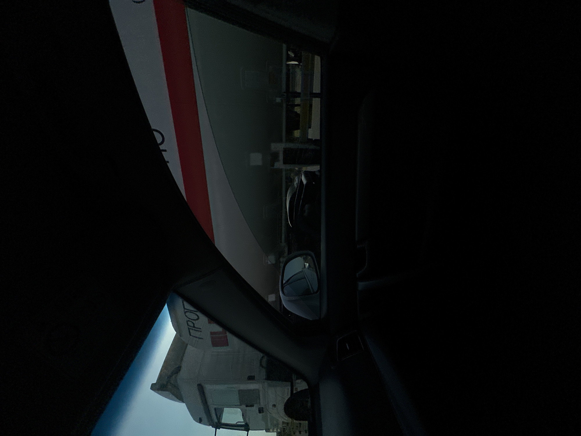 The image is taken from inside a vehicle, looking out at a large, white vehicle with red stripes. The perspective is tilted, suggesting the photo was taken in a landscape orientation or the vehicle is on an incline. The foreground is dominated by the dark interior of the car, with a side mirror visible. Reflected in the side mirror is a part of the interior of the car, and outside the window.
The large vehicle outside appears to be a truck or bus, with Cyrillic text visible on its side. Part of the text appears to be "ПРОГ". The scene seems to be outdoors during the daytime, given the bright sky visible. There are no people clearly visible in the image. The overall impression is one of being on a journey or observing a passing vehicle. The context of Tashtugizak, Uzbekistan, places the scene in a specific geographical location.
