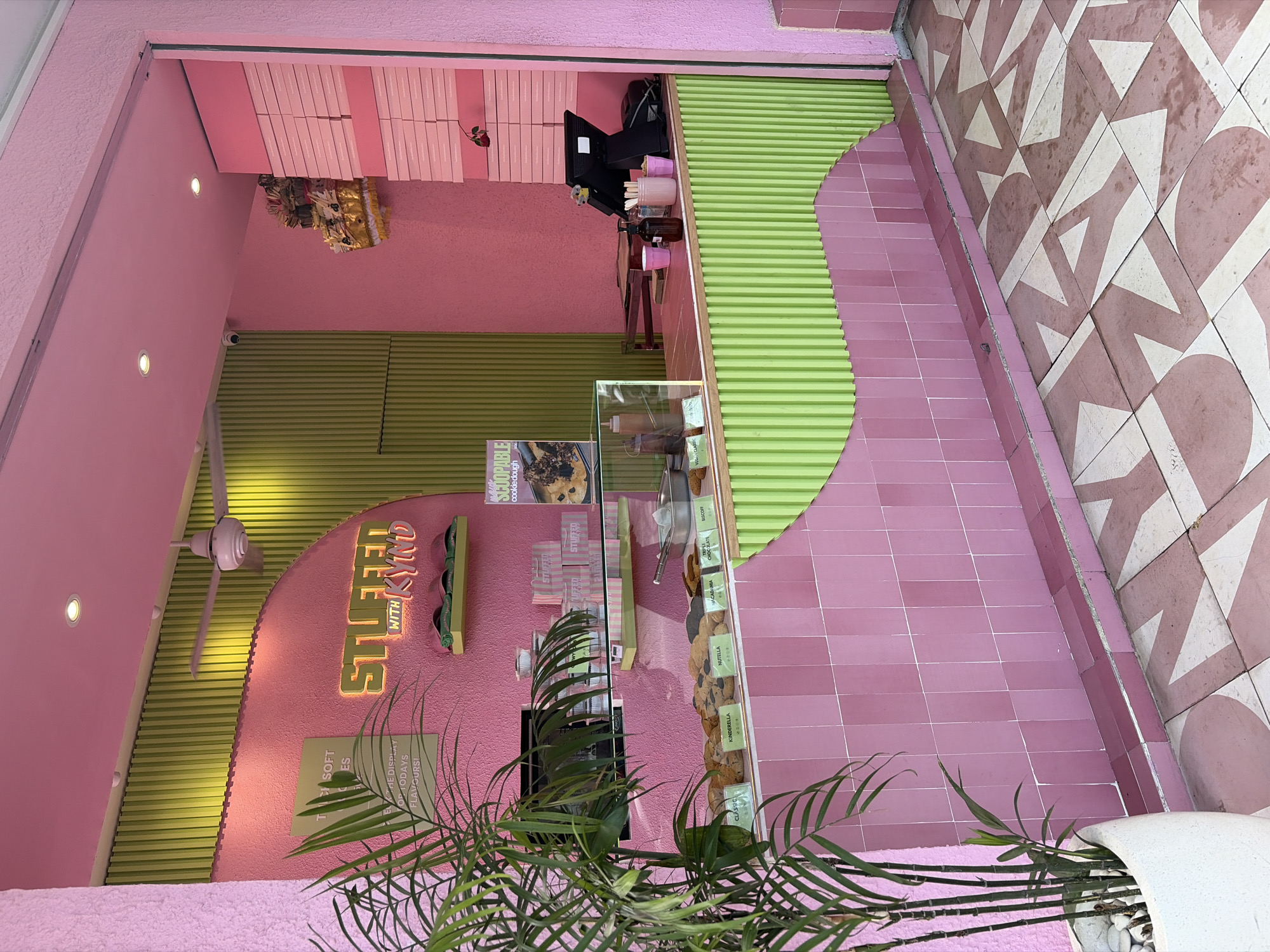 The image shows a bright pink and green dessert shop called "STUFFED WITH KIND" in North Kuta, Indonesia. The shop features a pastel color scheme, with pink walls, green corrugated metal accents, and pink tiled counters. A variety of cookies are displayed on the counter, each labeled with its flavor. Behind the counter, a cash register, a hand sanitizer dispenser, and pink cups are visible. A large plant in a white pot is positioned to the left of the counter. The shop's name is illuminated in neon green letters above the display case. A ceiling fan is mounted on the pink ceiling. In the background, a stack of pink boxes is visible on a shelf. The floor is tiled with a geometric pattern in pink and white. The overall impression is a whimsical and inviting dessert shop.