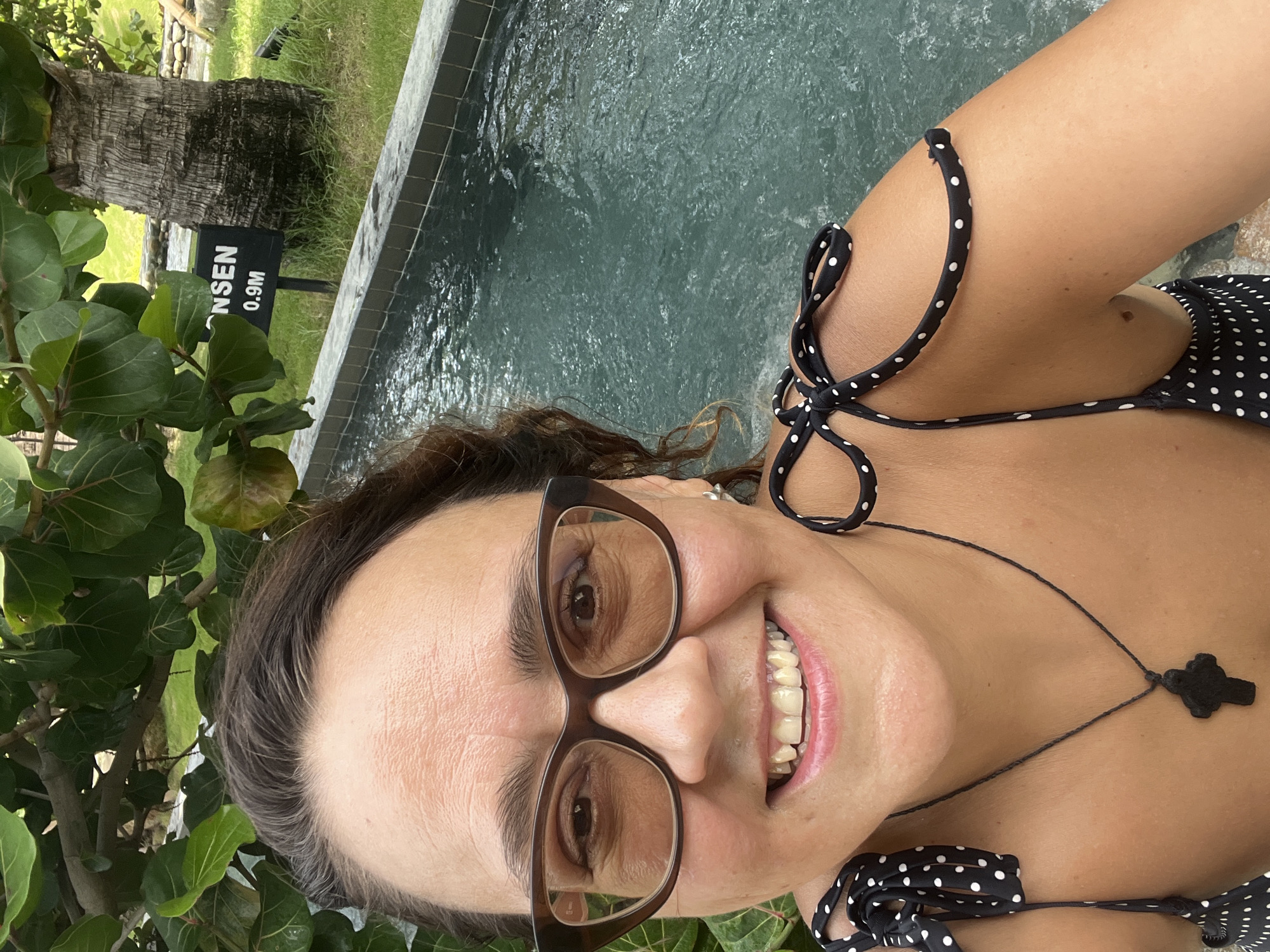 A woman with brown hair and glasses smiles while taking a selfie in a body of water. She is wearing a black and white polka dot bikini top with ties on the shoulders. A dark pendant necklace is visible around her neck. The background shows a pool with rippling water, green foliage, and a sign that reads "ONSEN 0.9M". The location is Nha Trang Ward, Vietnam.