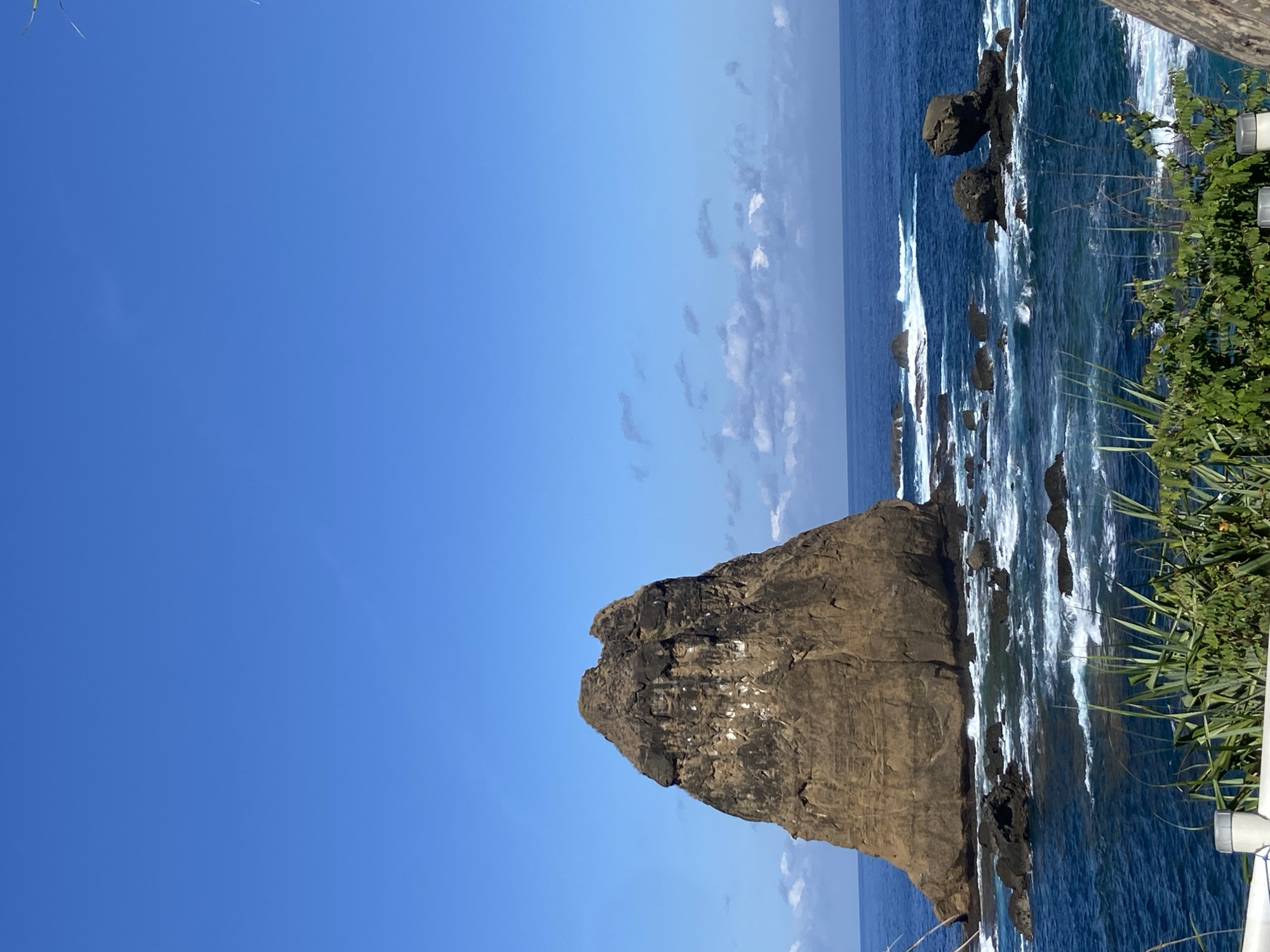 A landscape photograph shows a tall, brown rock formation jutting out of the sea, dominating the lower right quadrant. The rock formation appears weathered and textured, and it is partially covered in white streaks, possibly bird droppings. The dark blue sea surrounds the rock, with white waves crashing against its base and neighboring rocks. To the far right, green foliage grows near a white railing. The upper half of the image is filled with a clear, bright blue sky, with some wispy clouds along the horizon. The location is likely coastal, in Unknown, Indonesia.