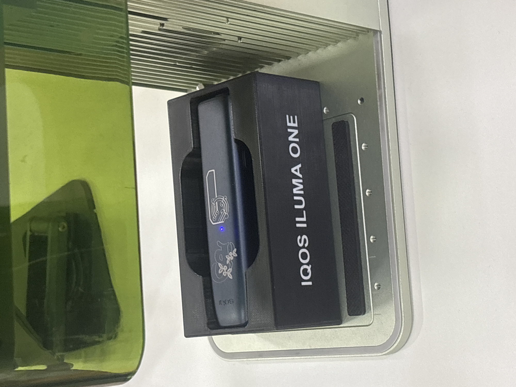 The image displays an indoor scene, potentially set in Tbilisi, Georgia, centered around an electronic device. A dark blue IQOS ILUMA ONE device is prominently featured, resting within a custom-made black holder. The device itself is adorned with intricate white or silver etchings, including a stylized animal, possibly a tiger or feline, near its upper section, and delicate floral designs, resembling cherry blossoms, towards the bottom. Below these patterns, the word "iqos" is subtly etched. A small, blue light emitted from the device indicates it is powered on or in the process of charging.
The black holder bears the text "IQOS ILUMA ONE" printed vertically in white capital letters on its right side. This holder is mounted on a brushed metallic panel, secured with visible screws, and attached to what appears to be a light-colored wall or vertical surface.
To the left of the mounted device, a large, translucent dark green object, likely a glass bottle or a decorative container, is partially visible. Its surface shows reflections of the surroundings, hinting at its material and form. Above the metallic panel and the green object, a metallic structure with horizontal slats or ribs can be seen, possibly part of a ventilation system, shelving, or a ceiling fixture. The background is predominantly a plain white or light-colored wall, providing a clean and understated setting for the items. The overall impression is that of an organized and functional display of a personal electronic device.