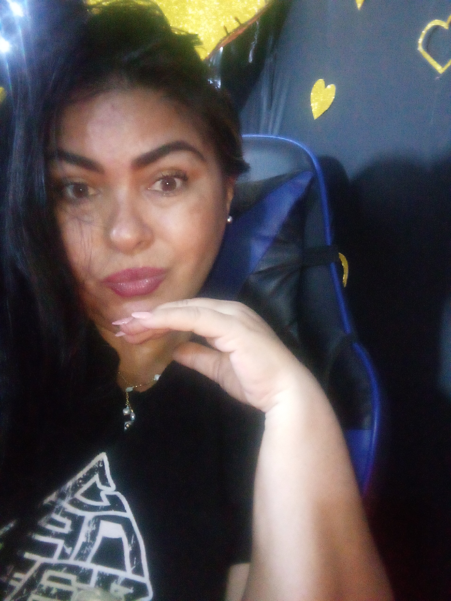 A woman with dark hair and light skin is in a selfie, wearing a black shirt and a necklace with a pendant. She is resting her hand on her chin. She is seated in a black and blue gaming chair. The background is a dark surface decorated with yellow hearts. The location, Juan Griego, Venezuela, is not discernible from the image.
