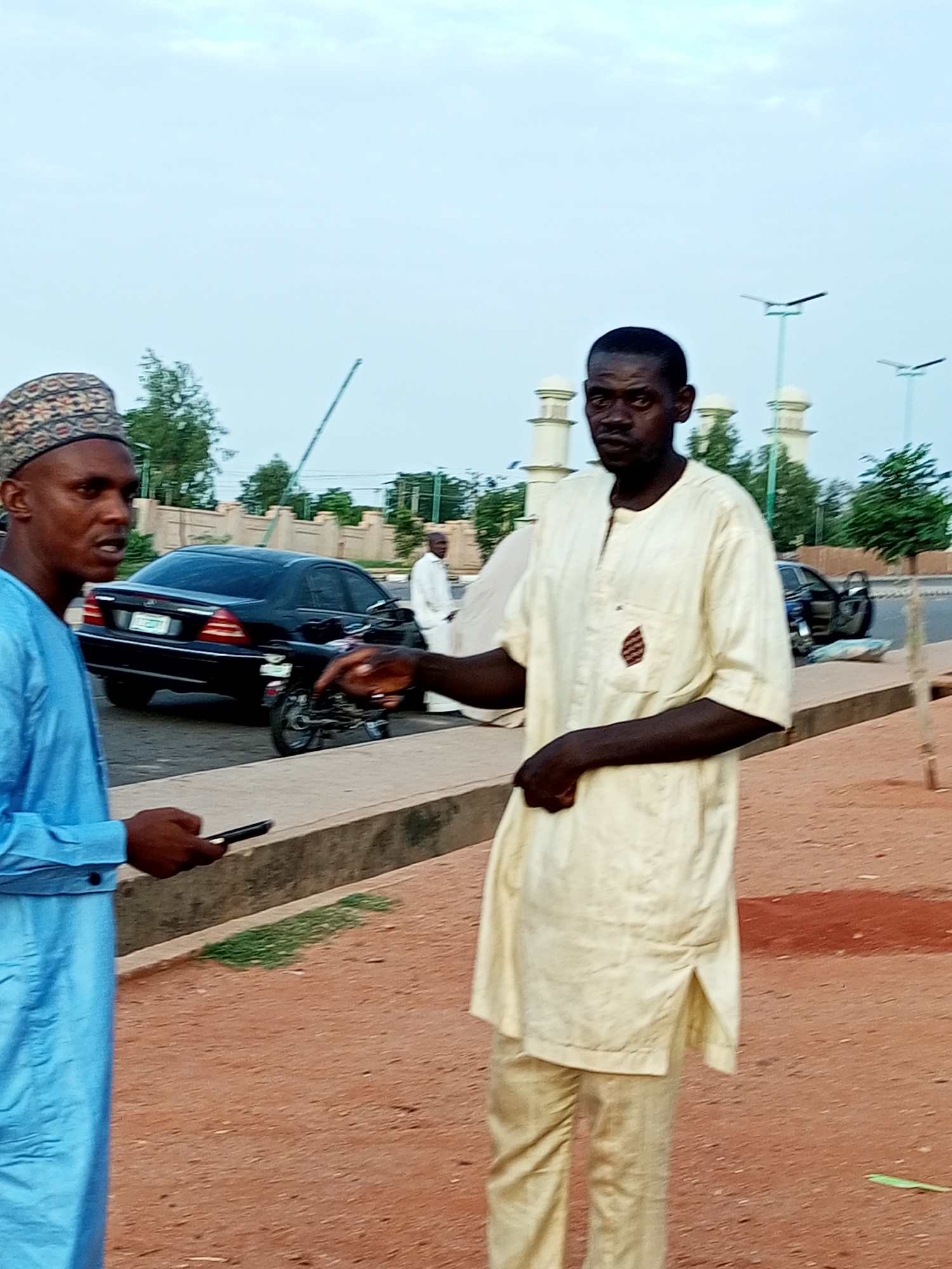 Two men are interacting on a dusty street in the city of Gombe, Nigeria. The man on the left is wearing a blue traditional robe and a patterned cap, holding a smartphone in his left hand. The man on the right is wearing a light-colored traditional outfit, looking directly at the camera. In the background, a black car and a motorcycle are parked on the side of the road. Several lampposts and architectural elements that appear to be part of a mosque or similar structure are visible in the distance. The ground is covered in reddish-brown dirt, with some patches of dry grass. The sky is overcast with light clouds.