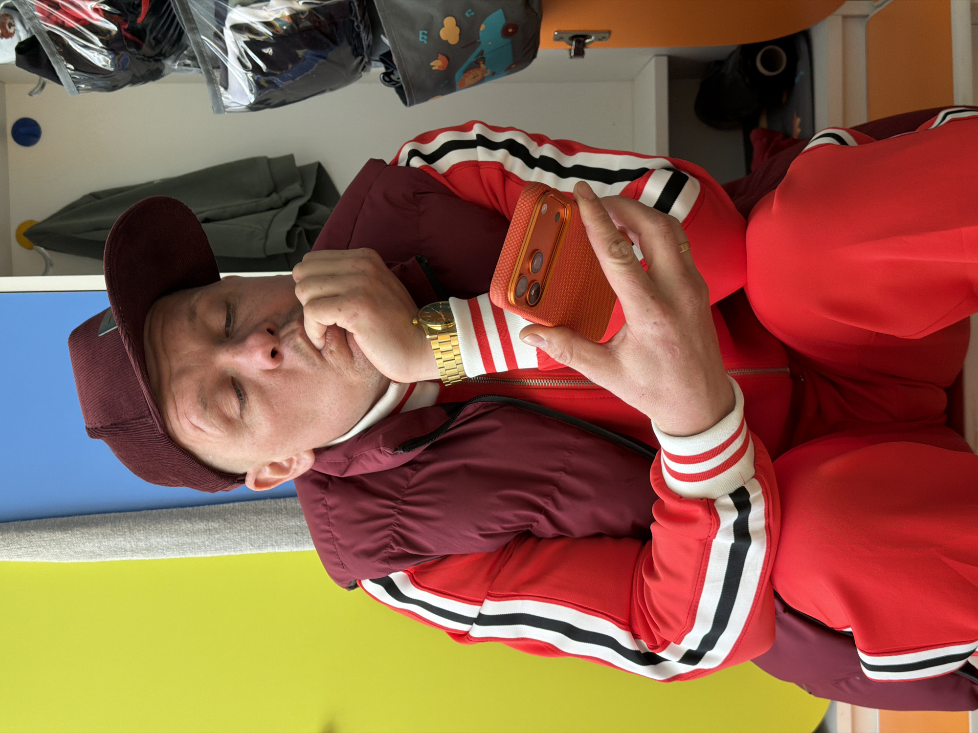 A man is seated in front of a closet with an open drawer, holding an orange cell phone in his right hand. He's wearing a maroon corduroy baseball cap, a white turtleneck, a red tracksuit with white stripes bordered in black down the sleeves and legs, and a dark burgundy puffer vest. His left hand is clenched near his face. He's wearing a gold watch on his left wrist and a gold band on his right ring finger. The open closet drawer is beneath him, and its back wall is painted blue and its bottom is painted yellow. In the top left corner of the image, clothing items hang on hangers within the closet. A black cylindrical object is visible inside the closet above the man's right shoulder. The overall environment suggests an interior space, likely a bedroom or dressing room. There is no indication of the location in Chaikovsky, Russia.