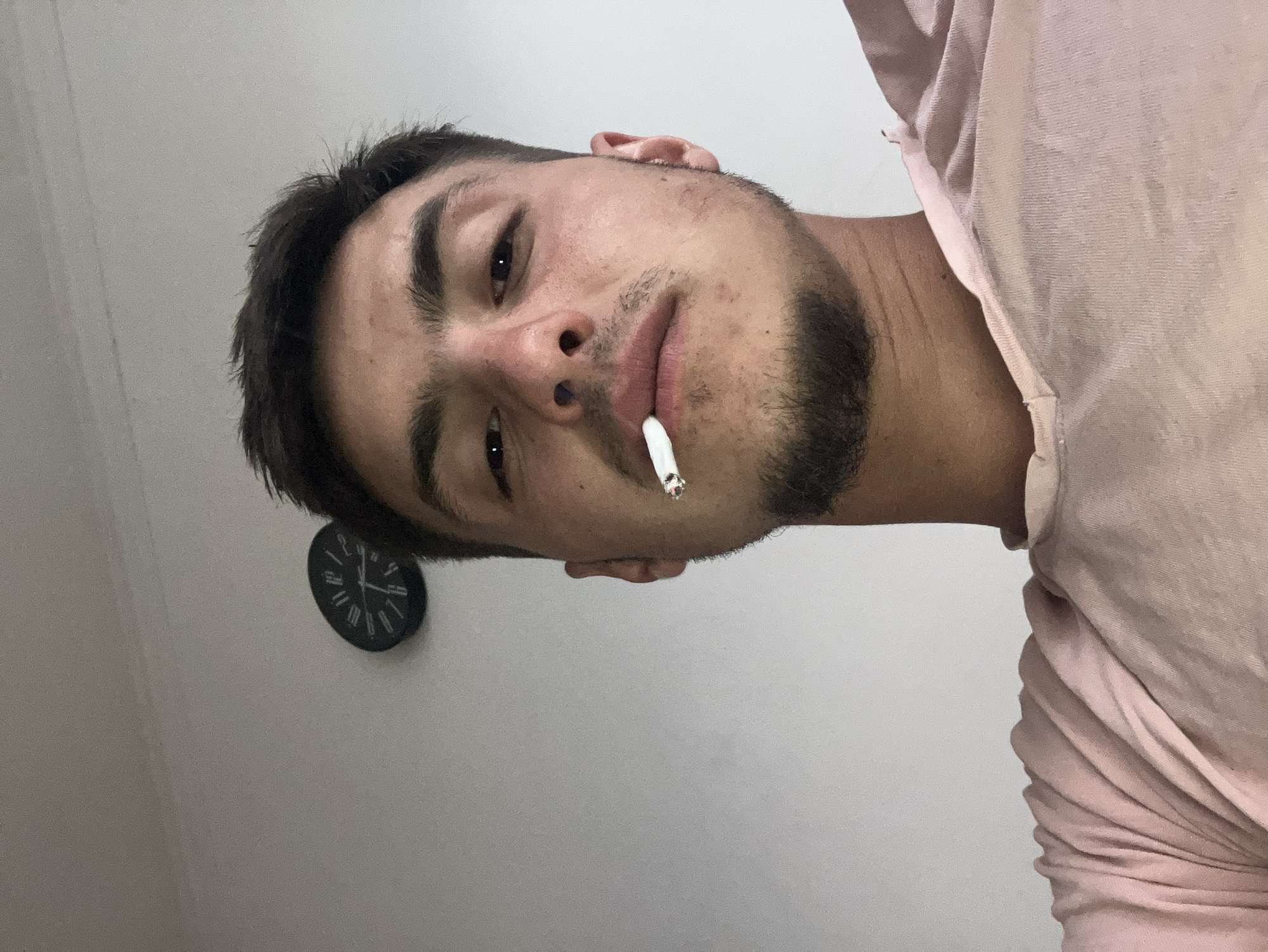 A man with dark hair and a short beard is holding a lit cigarette in his mouth. He is wearing a pink shirt. A clock is visible on the wall behind him. It is not possible to determine the location in the city of Plovdiv, Bulgaria from the image alone.