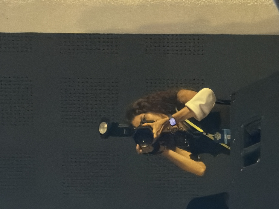 An indoor scene in the city of Unknown, Indonesia, features a female individual actively operating a professional camera. The woman, with long, dark, curly hair, is wearing a dark sleeveless top, a yellow lanyard with an identification badge, a smartwatch on her left wrist, and rings on her right hand. Her right forearm is partially covered by a white sleeve. She holds a professional-grade camera equipped with a large lens and an external flash unit mounted on its hot shoe, positioned forward. The camera is raised to her eye level, indicating an active photographic engagement. In the immediate foreground, the top of a dark, angular structure, possibly a stage monitor or speaker, is visible. The background consists of a dark, perforated acoustic wall to the left, and a lighter, textured vertical surface on the right. The overall illumination of the scene is dim, consistent with an indoor event or performance setting.