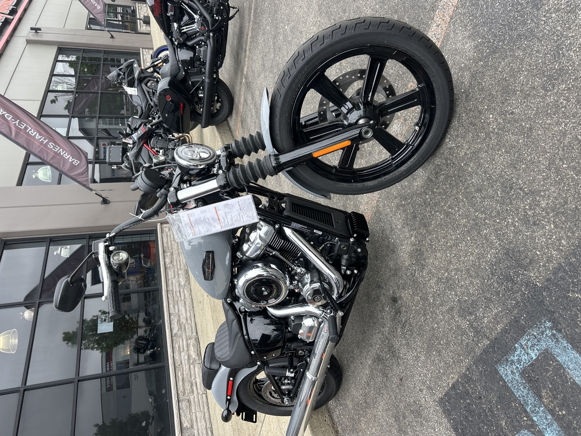 This is a medium-shot, eye-level, full-color photograph of a gray Harley-Davidson motorcycle parked on an asphalt surface outside a dealership. The motorcycle is the primary subject and is angled slightly towards the viewer's left. It is a street bike with a black seat, black wheels with silver spokes, and chrome accents on the engine and exhaust. A price tag is attached to the handlebars.
In the background, a building with large windows and a stone facade is visible. A banner with the text "BARNES HARLEY-DAVIDSON" is hanging from the building. Other motorcycles are parked in front of the building, some of which are black. The sky is overcast, suggesting a cloudy day. The ground in the foreground has faint blue markings that appear to be part of a parking lot designation. The scene is set in a commercial area, likely a motorcycle dealership in the Township of Langley, Canada.