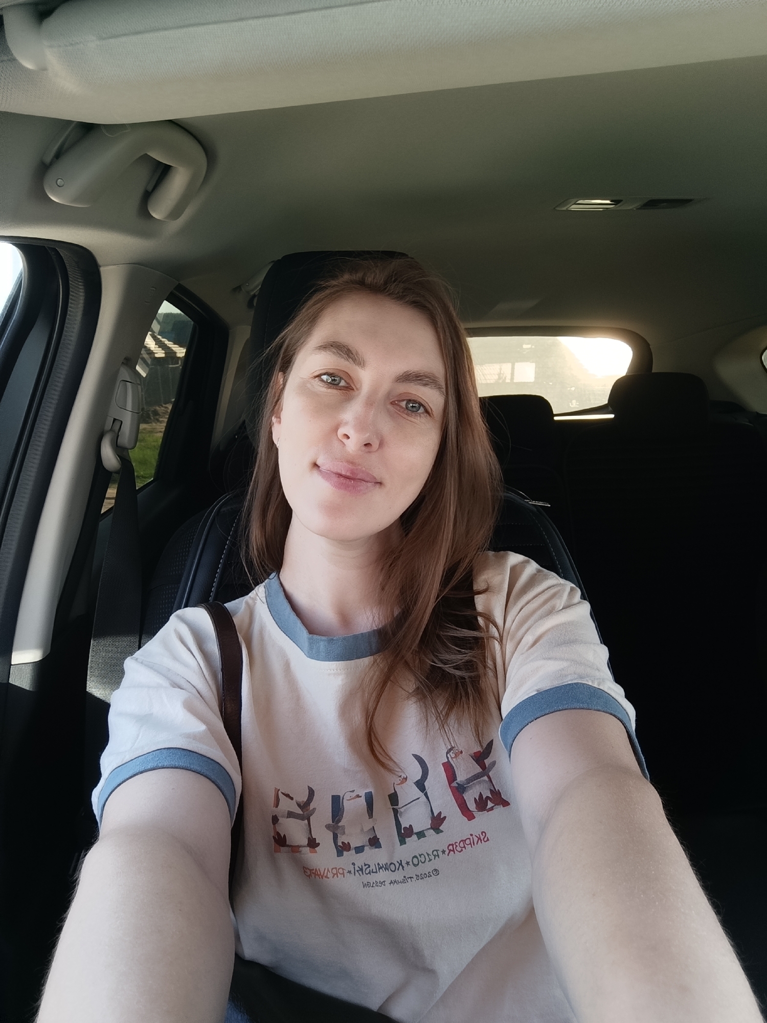 A woman is taking a selfie while seated in the driver's seat of a car. She has light skin, brown hair, and is wearing a light-colored t-shirt with a blue collar and an indecipherable pattern. The car's interior is visible, including the dashboard, passenger seat headrest, and a seatbelt across her torso. Light is entering through the back window of the car. The location within the city of Chaikovsky, Russia, cannot be determined from the provided image.