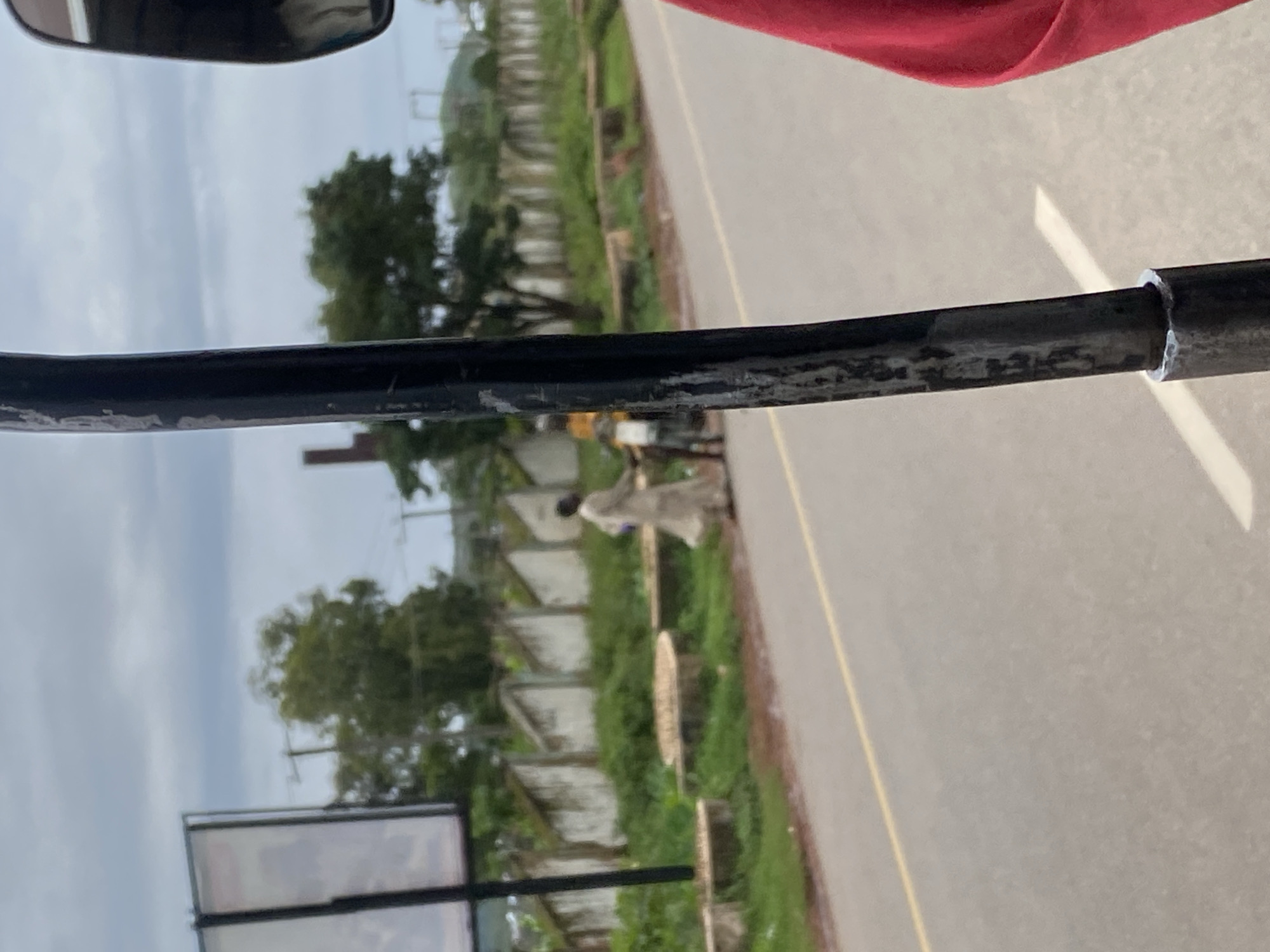 The image shows a scene on a road in Rafin Par, Nigeria. In the foreground, a vertical black metal pole, possibly part of a vehicle, is visible. To the right, a portion of a person's red garment is present, suggesting the photographer is within a vehicle. In the mid-ground, two figures are walking on the side of the road. One figure is wearing a long, light-colored garment, and the other is wearing darker clothing and appears to be on a motorbike. A road with a yellow line is present, separating the immediate foreground from the background. The background features a wall with openings, overgrown green vegetation, trees, and a cloudy sky. In the distance, industrial structures and hills are faintly visible.