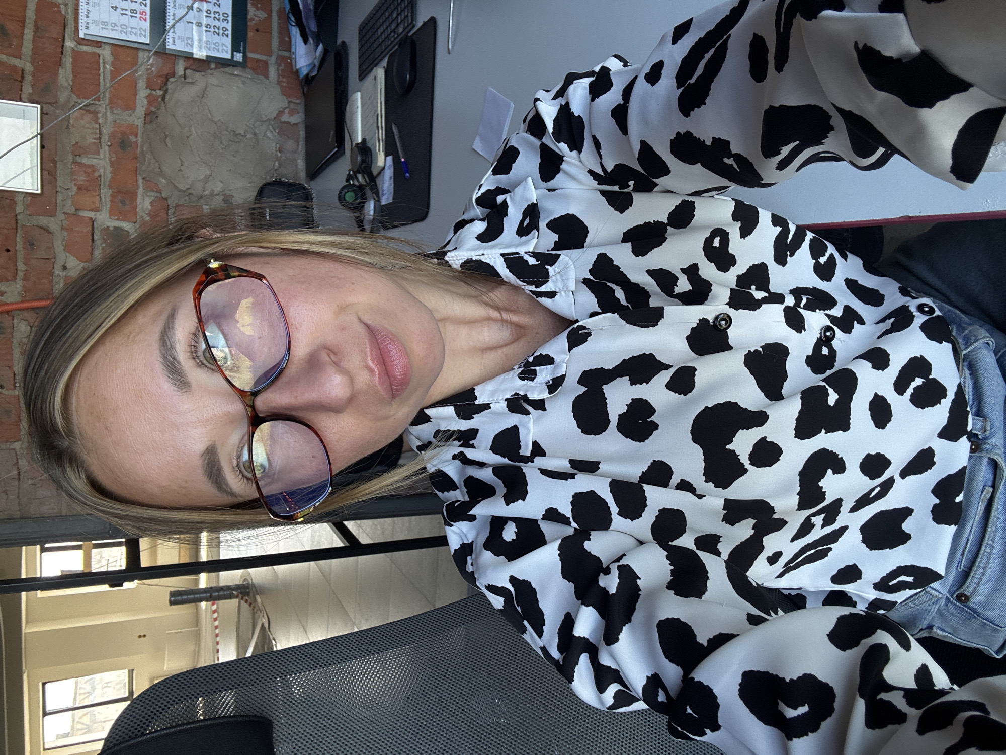 In the city of Chaikovsky, Russia, a woman with blonde hair and glasses is sitting at a desk, taking a selfie. She is wearing a black and white spotted shirt and blue jeans. The background shows a construction site, with exposed brick walls and windows. There is also a computer and a calendar on the desk. The lighting in the image is bright and natural, suggesting it was taken during the day. The woman's facial expression is playful, with her lips pursed.