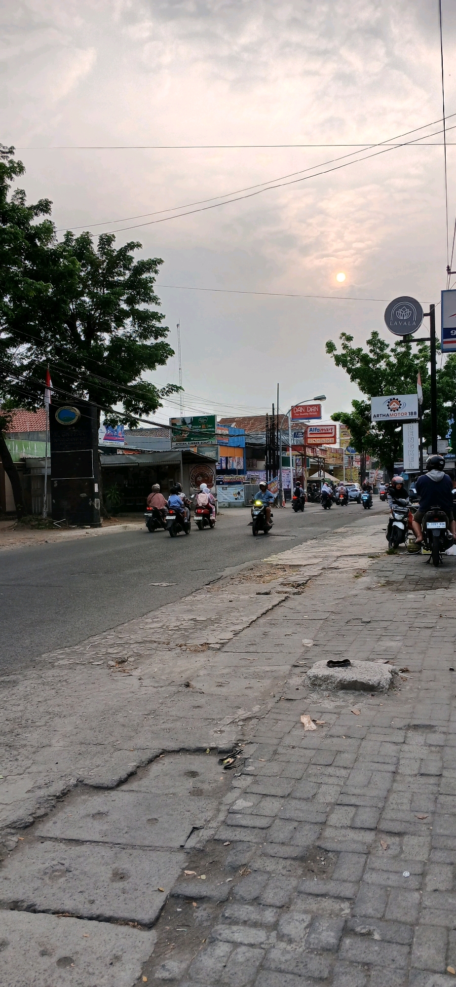 The image depicts a street scene in Cirebon, Indonesia, likely during daylight hours with overcast skies and the sun visible as a bright orb. The street is paved with asphalt and concrete blocks, showing signs of wear and tear with numerous cracks and potholes. A row of buildings with various storefronts lines the right side of the street, including signage for "DAN DAN," "Alfamart," "LAVALA," and "ARTHAMOTOR 188." The left side of the street features a large tree, a dark monument with a circular emblem, and flags. A stream of motorcycles is actively navigating the street, indicating traffic flow. Several individuals are riding these motorcycles, some with passengers, all appearing to be engaged in their commute. Power lines are visible overhead, strung between poles. The overall impression is of a typical urban street environment in Indonesia.