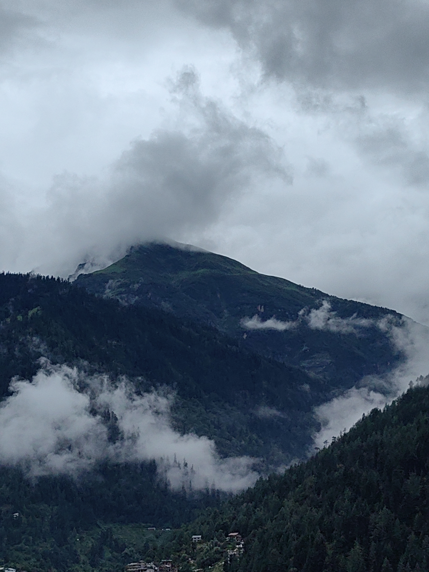 A vertical, low-angle shot captures a cloudy, overcast day in Vashisht, India, with a focus on the mountainous landscape. Dark, coniferous trees densely cover the steep slopes of the mountains. Wisps of white fog and clouds drift through the valleys and cling to the mountainsides, partially obscuring the view. A single, prominent peak rises in the upper center of the frame, its upper slopes covered in a vibrant green, hinting at vegetation above the treeline. The sky above is filled with heavy, gray clouds, contributing to the moody and dramatic atmosphere of the scene. Small buildings are visible in the distance, nestled at the base of the mountains, suggesting a human presence within this natural environment. The overall impression is one of a remote and rugged mountain region experiencing a day of persistent cloud cover.