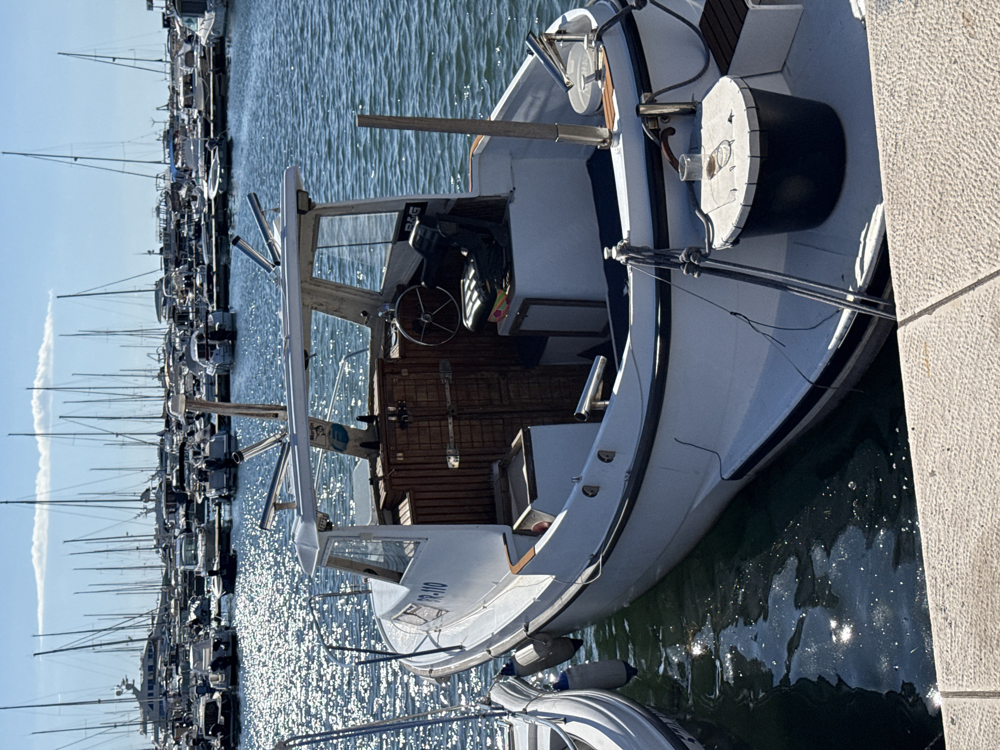 A white motorboat with the registration number BR-110 is docked in a marina in Bar, Montenegro. The boat is positioned in the foreground, with its bow pointing towards the right side of the image. It has a covered cabin with windows and a steering wheel visible inside. The boat is equipped with fishing rod holders and fenders. In the background, numerous sailboats and other vessels are moored in the marina under a bright blue sky with some clouds. The water is calm, reflecting the sunlight. The image captures a typical scene in a Mediterranean marina.
