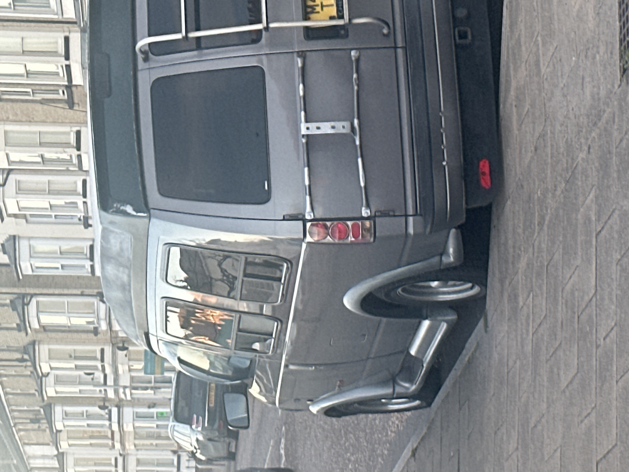 The image captures a close-up of the rear of a gray van parked on a street in East Suffolk, United Kingdom. The van has a ladder attached to the back and a rear window. The scene includes a sidewalk with a patterned surface adjacent to the parked van. In the background, a building with multiple windows is visible, suggesting an urban or residential area within the city. Another vehicle is partially visible behind the main van. No people are visible in the frame.