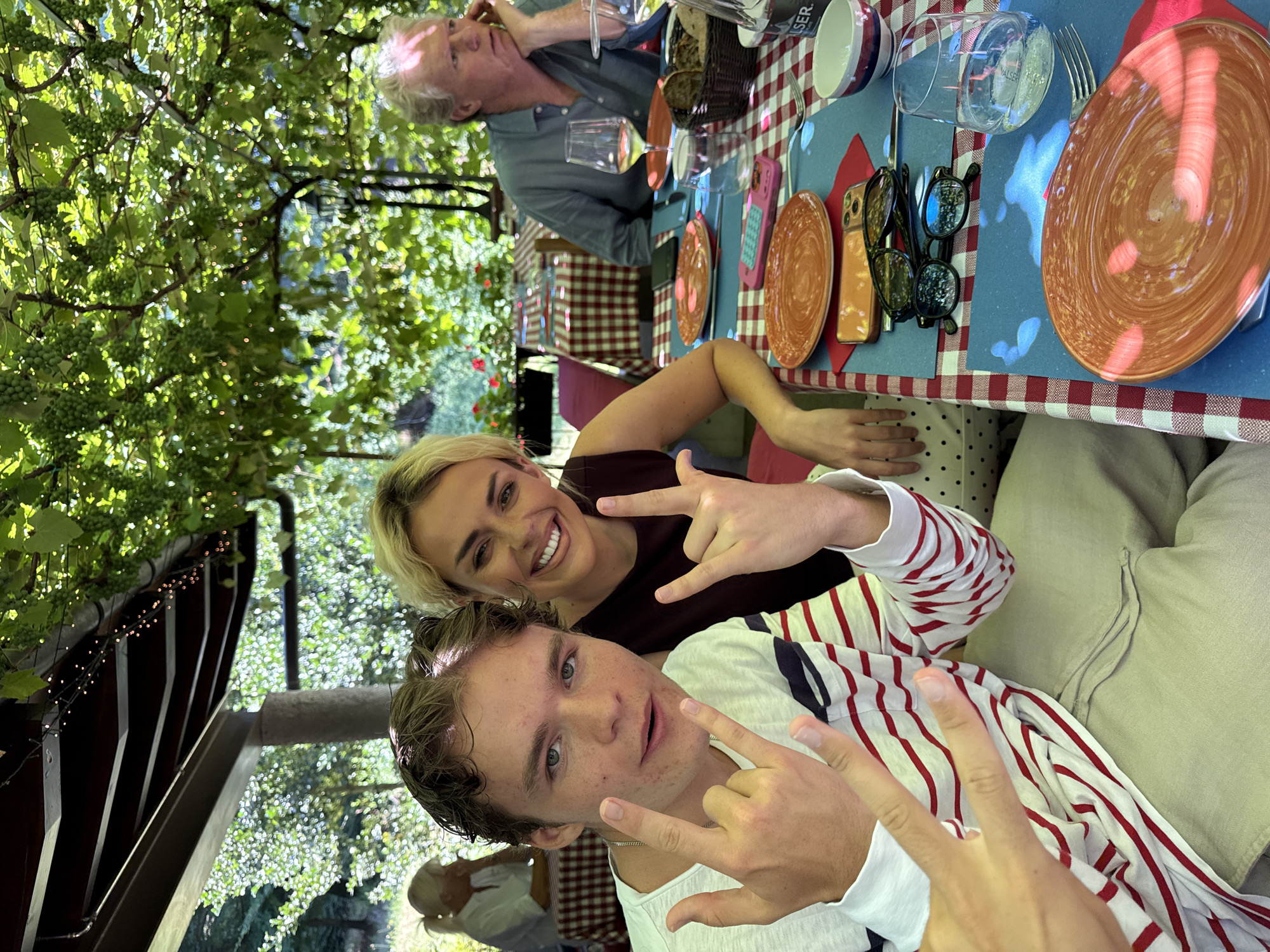 A medium shot captures three individuals at an outdoor table with a checkered tablecloth. The table is set with orange plates, glasses, and a basket of bread. Overhead, grapevines with green grapes cast dappled light on the scene. In the foreground, a young man with curly brown hair grins at the camera, holding up his hands in a rock-and-roll gesture. He wears a striped shirt and light-colored pants. To his right, a woman with blonde hair smiles broadly, also making a similar hand gesture. Further back, an older man with gray hair rests his chin on his hand, looking to the side. The location is Agno, Switzerland.