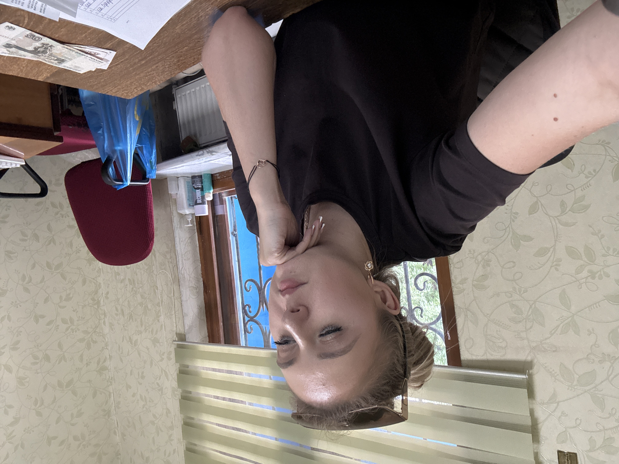 A young woman with blonde hair tied up and sunglasses on her head sits at a wooden desk, looking down with her chin resting on her hand. She is wearing a black t-shirt and a bracelet with a heart charm. In the background, there is a window with blinds, a red chair with a blue plastic bag hanging from it, and a patterned wallpaper. On the desk, there are some banknotes, papers, and bottles of cleaning supplies. The scene appears to be an office or a home workspace.