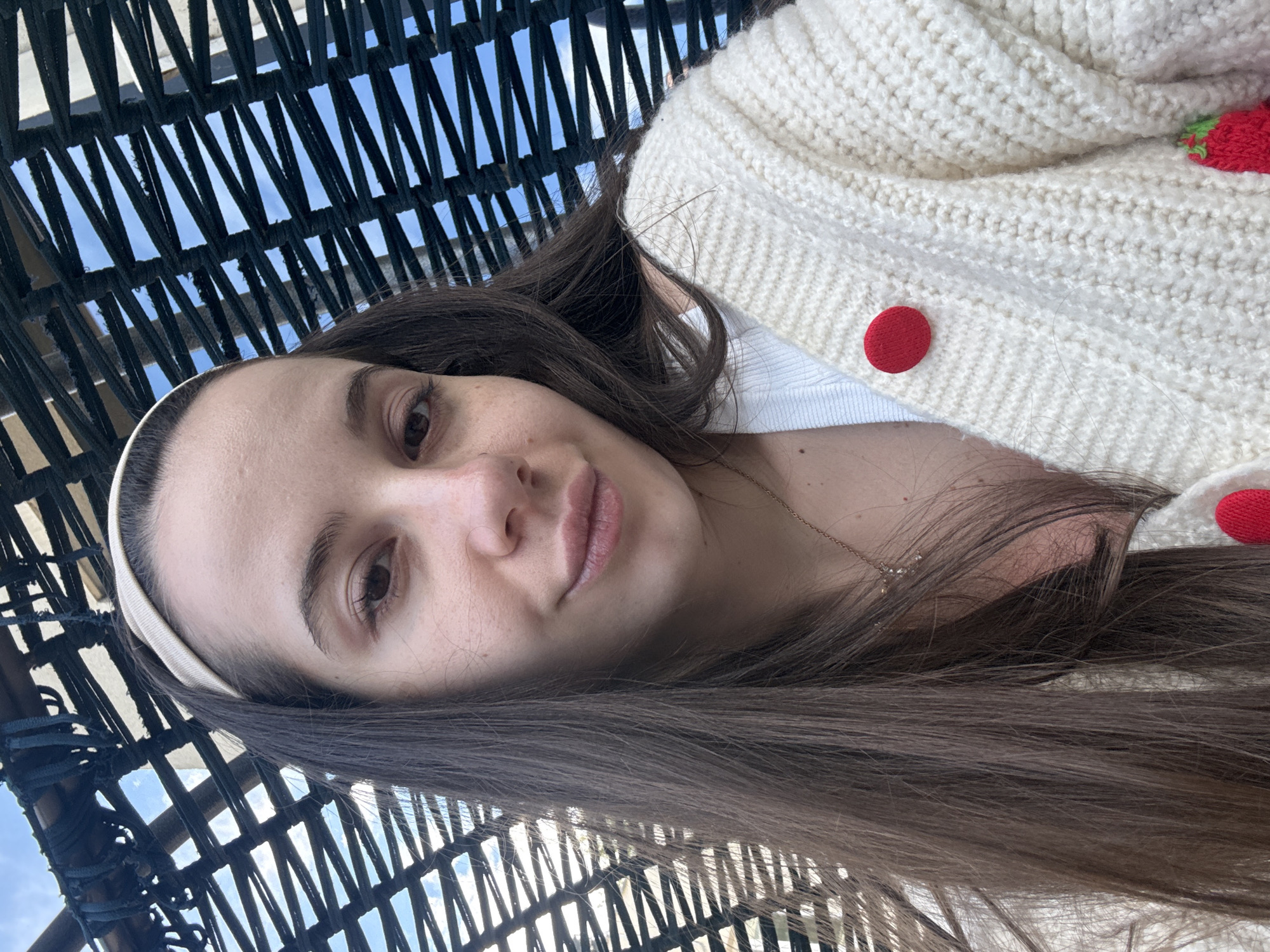A woman with brown hair and a white headband is taking a selfie. She is wearing a white cardigan with red buttons and a white top. She is sitting in a wicker chair with a black frame. The sky is visible through the wicker weave. The location is indeterminate, and there is no indication of Varna, Bulgaria.