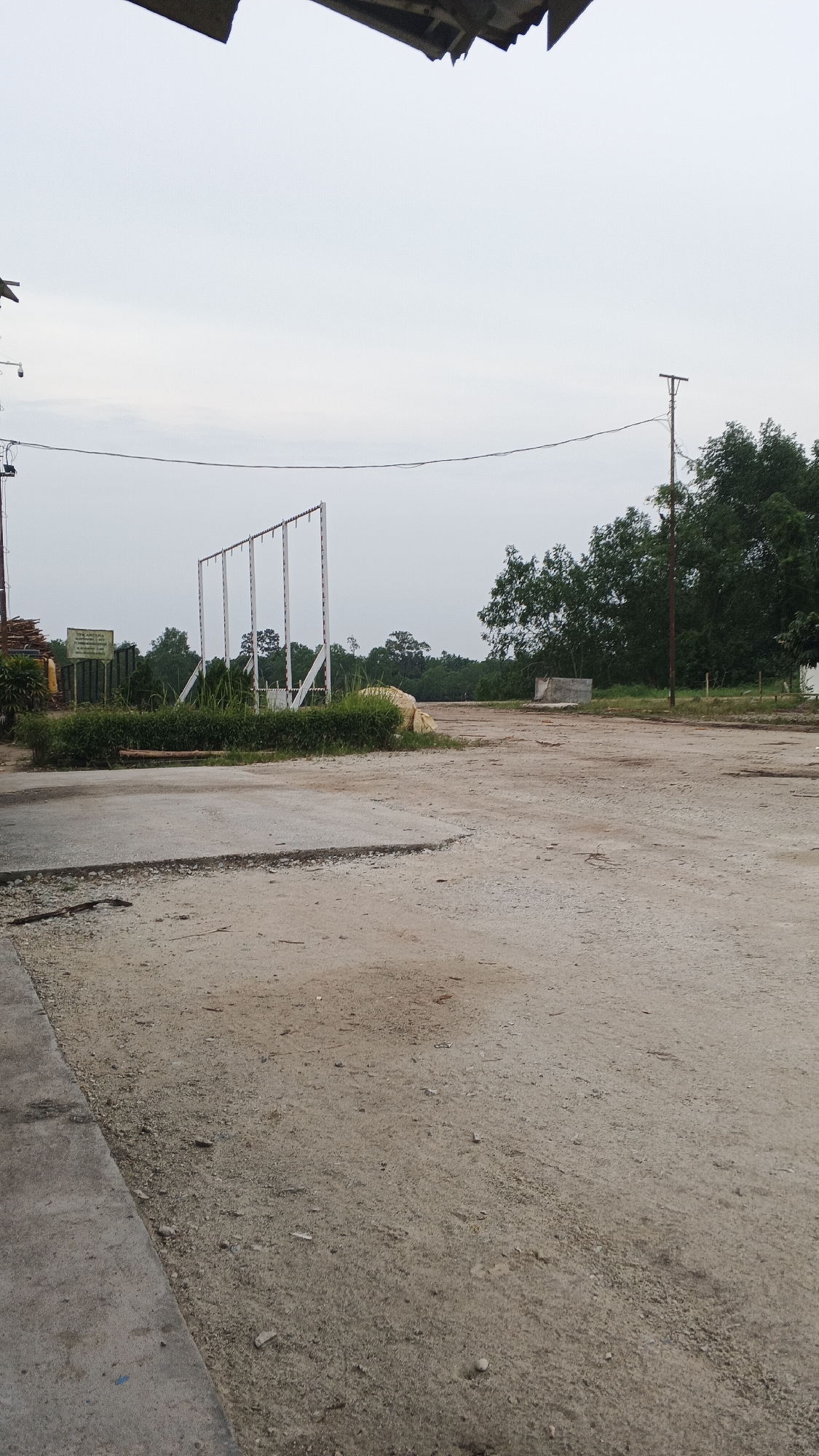 The image depicts an outdoor scene in Merempan Hilir, Indonesia, characterized by a dusty, unpaved ground surface. A concrete structure is visible in the foreground on the left, with a dirt pathway extending into the mid-ground. In the background, a row of tall, white, vertical posts with horizontal crossbars stands in a line, suggesting a framework for some purpose, possibly for a fence or a support structure. Adjacent to this structure is a patch of dense green vegetation, bordered by a larger pile of what appears to be sand or material wrapped in a light-colored tarp. Further in the background, mature trees form a dense green backdrop against a hazy sky. A utility pole with wires stretches across the scene. The overall impression is of an undeveloped or industrial outdoor area.