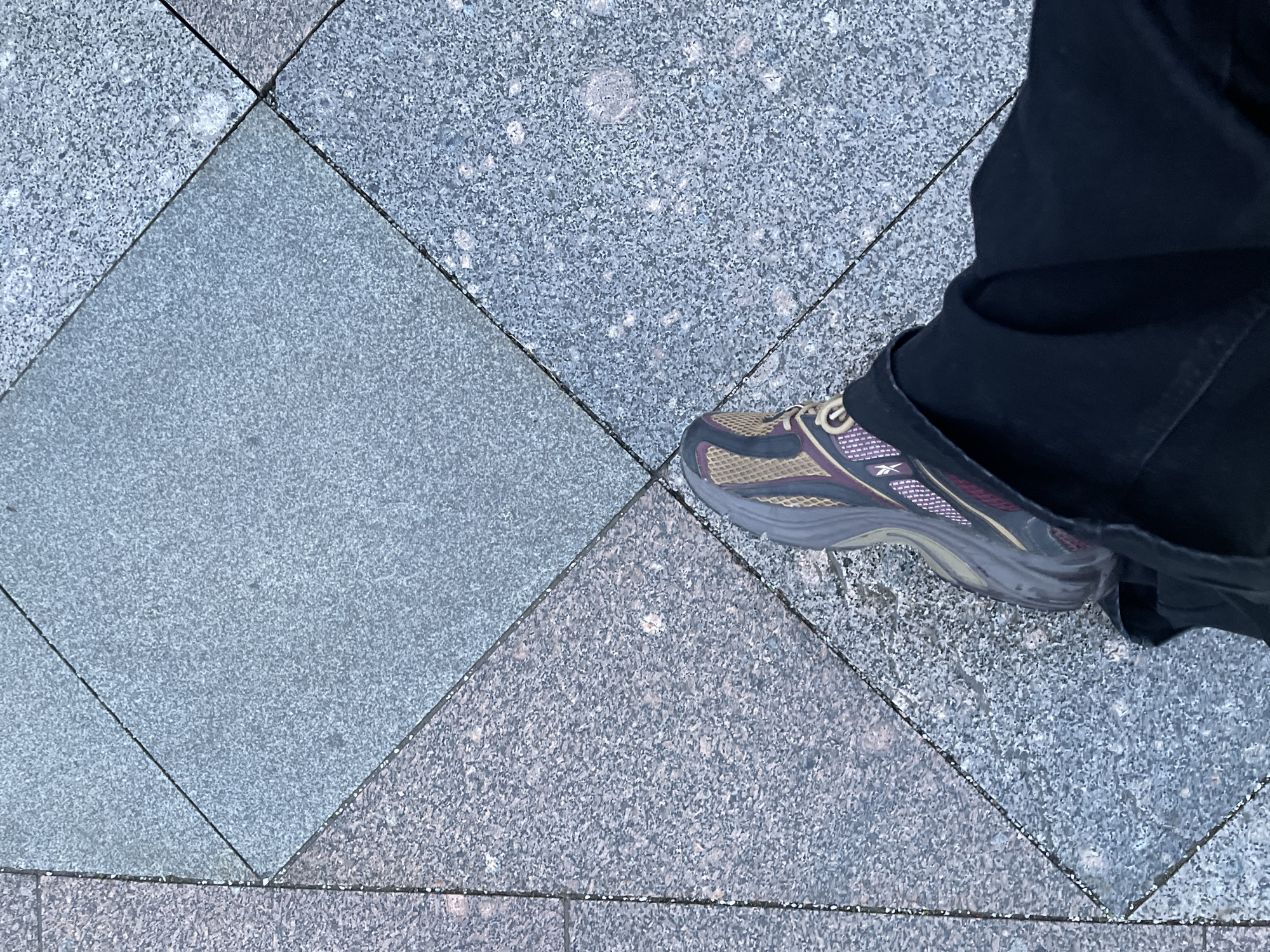The image shows a foot wearing a Reebok shoe on a paved surface. The paving consists of square tiles laid diagonally. The tiles are of two different shades of grey. The person is wearing dark-colored pants. The background is out of focus, not allowing determination of the location in Moscow, Russia.