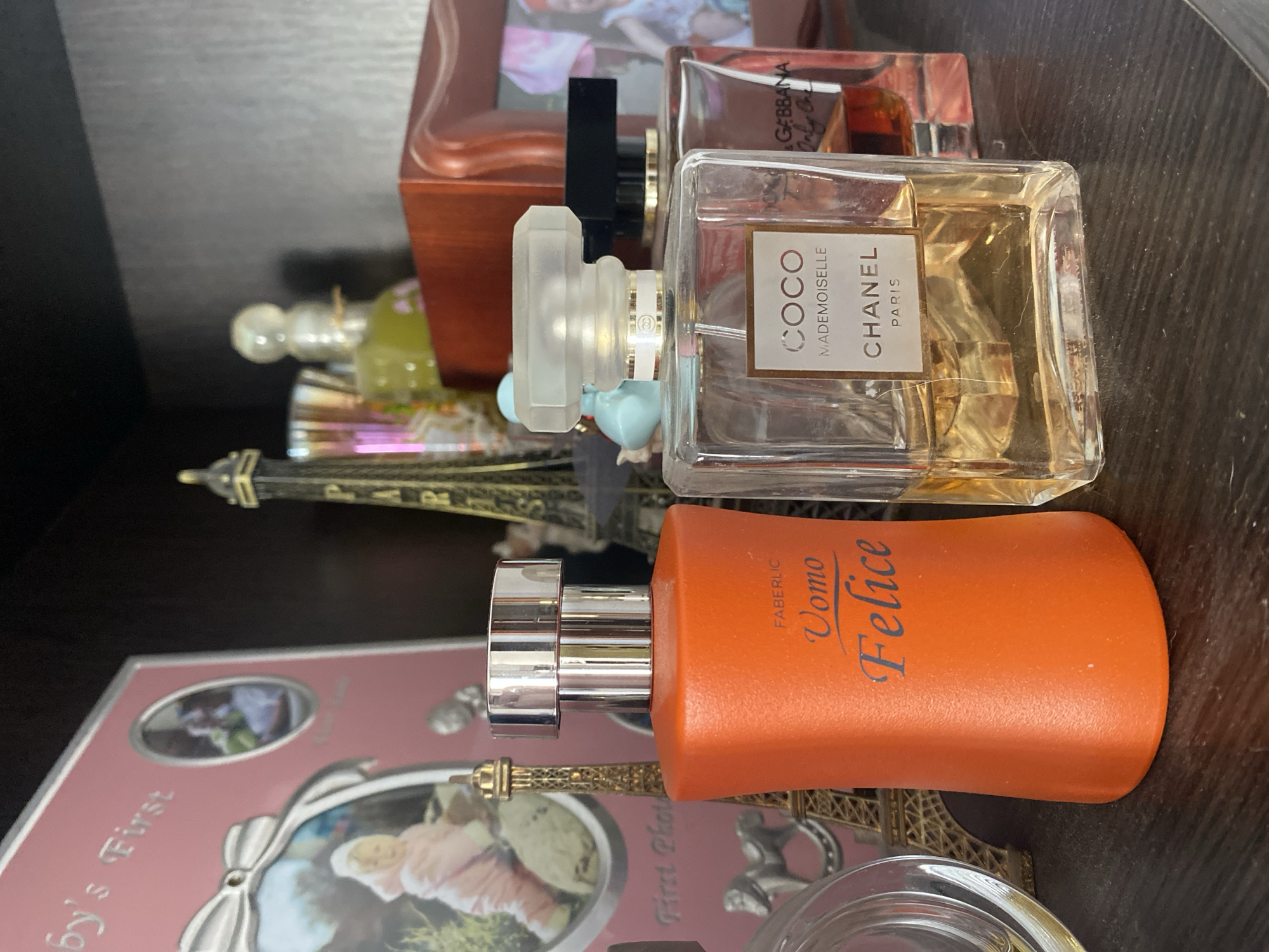 The image depicts a close-up, eye-level shot of a shelf displaying several perfume bottles and decorative items. The foreground is dominated by an orange-colored perfume bottle labeled "FABERLIC Uomo Felice". To its right is a clear glass bottle of "CHANEL COCO MADEMOISELLE" perfume. Behind these, partially visible, are other perfume bottles and decorative Eiffel Tower figurines. In the background, a pink picture frame with a baby's photo and the text "baby's First Photo" is also present. The shelf is dark wood, and the overall lighting suggests an indoor setting. No people are visible in the image. The location in Chornomorsk, Ukraine, is inferred from external information and is not visually represented within the image itself.