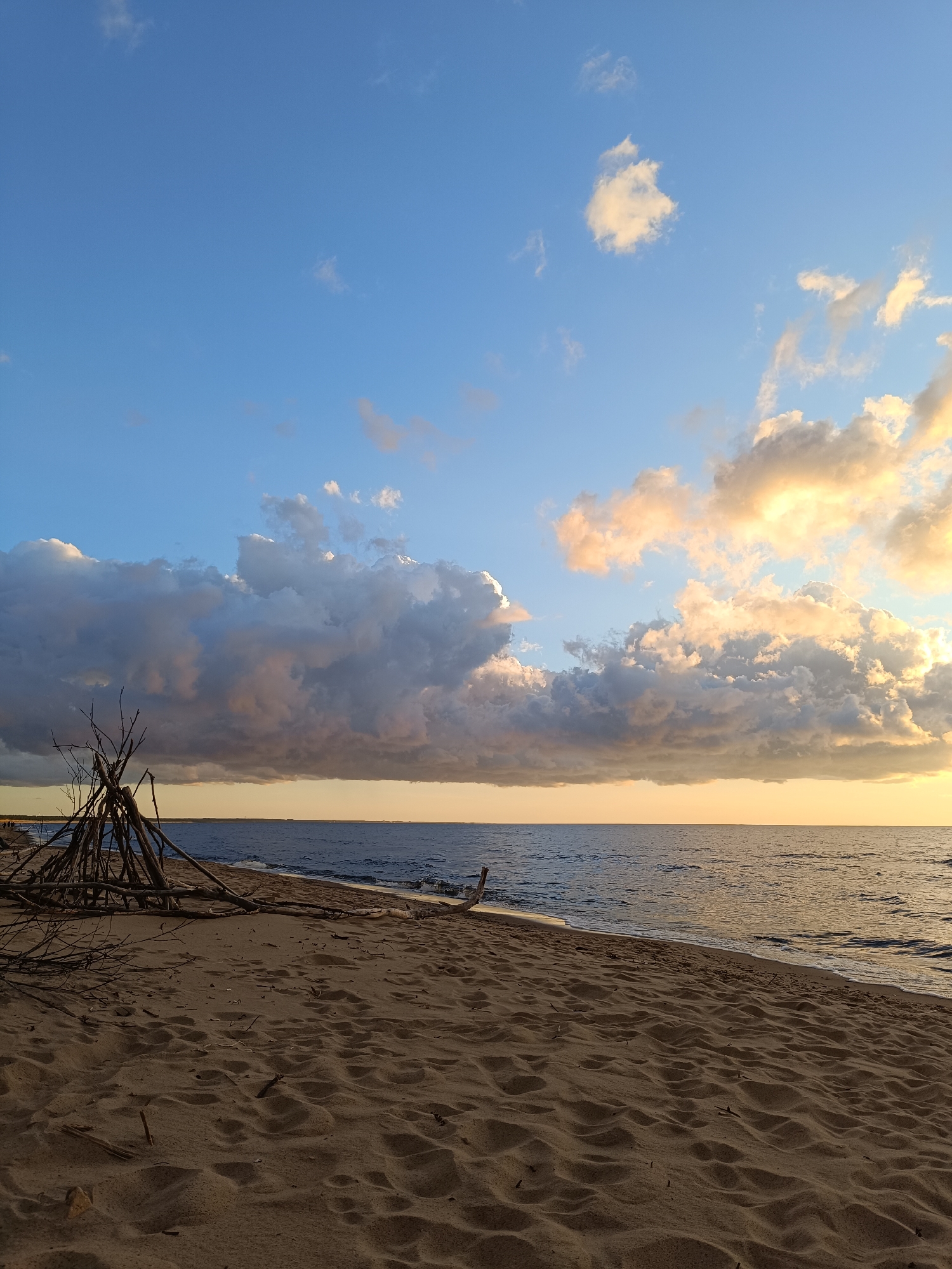 The image shows a sandy beach with the ocean in the background under a cloudy sky. In the foreground, a makeshift shelter made of sticks and branches is constructed on the sand. The ocean extends to the horizon where it meets a sky filled with dramatic clouds illuminated by the setting sun. The sand is textured with footprints and small debris. A few people are visible in the distance along the shoreline. The overall scene depicts a natural landscape at dusk.